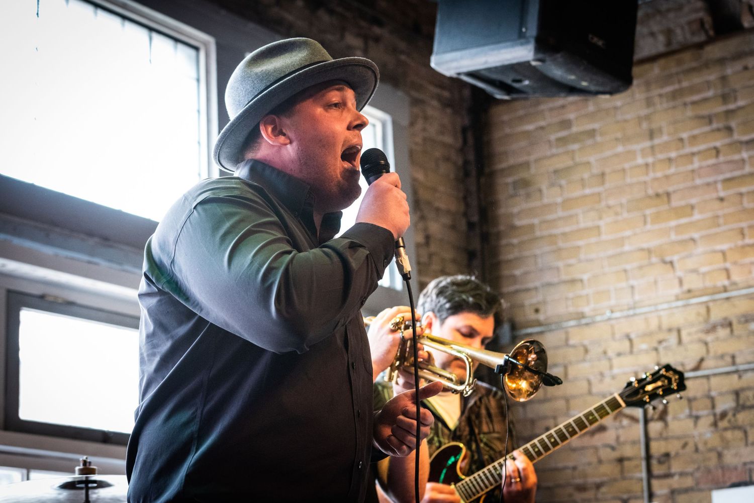 A performer singing into a microphone at a venue with brick walls and musicians playing behind them.