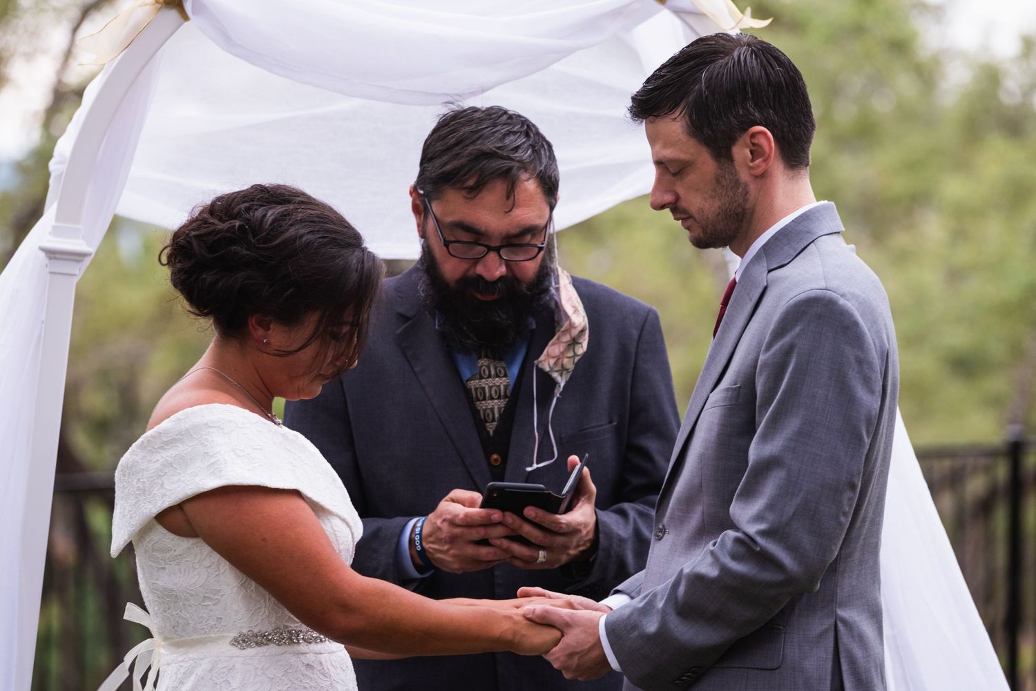 Wedding ceremony under white arch with officiant reading from device.