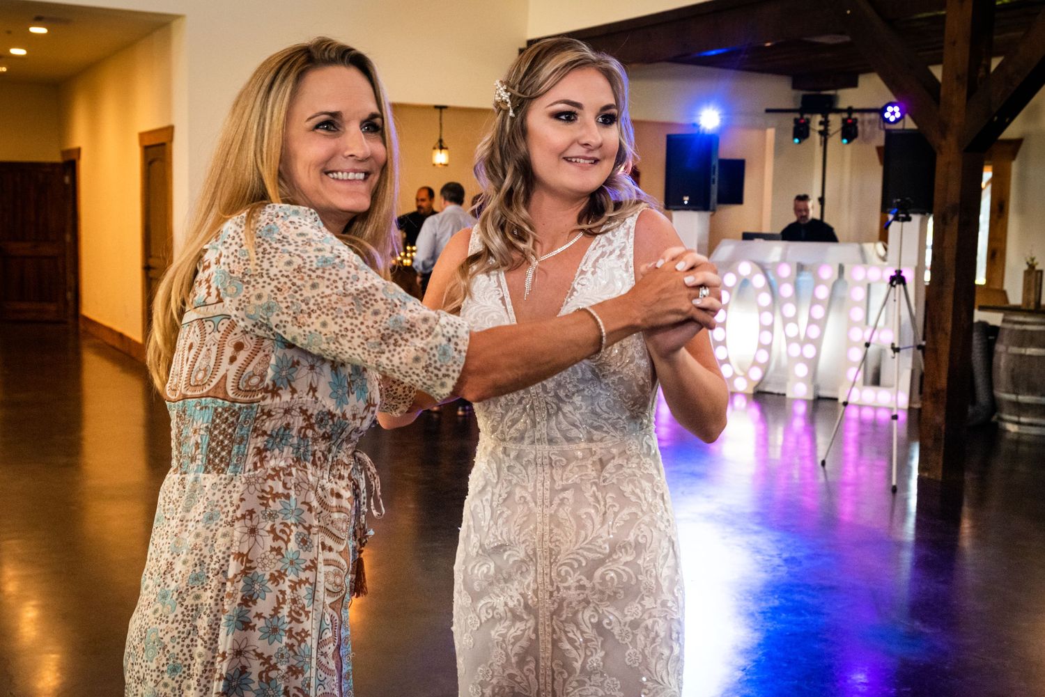 Two women in elegant dresses dance together at a reception with illuminated decor in the background.