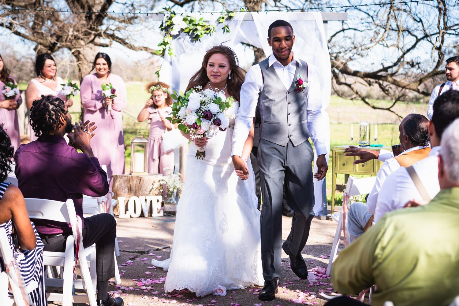 Happy newlyweds walk down the aisle after outdoor wedding ceremony with guests and bridesmaids in background.