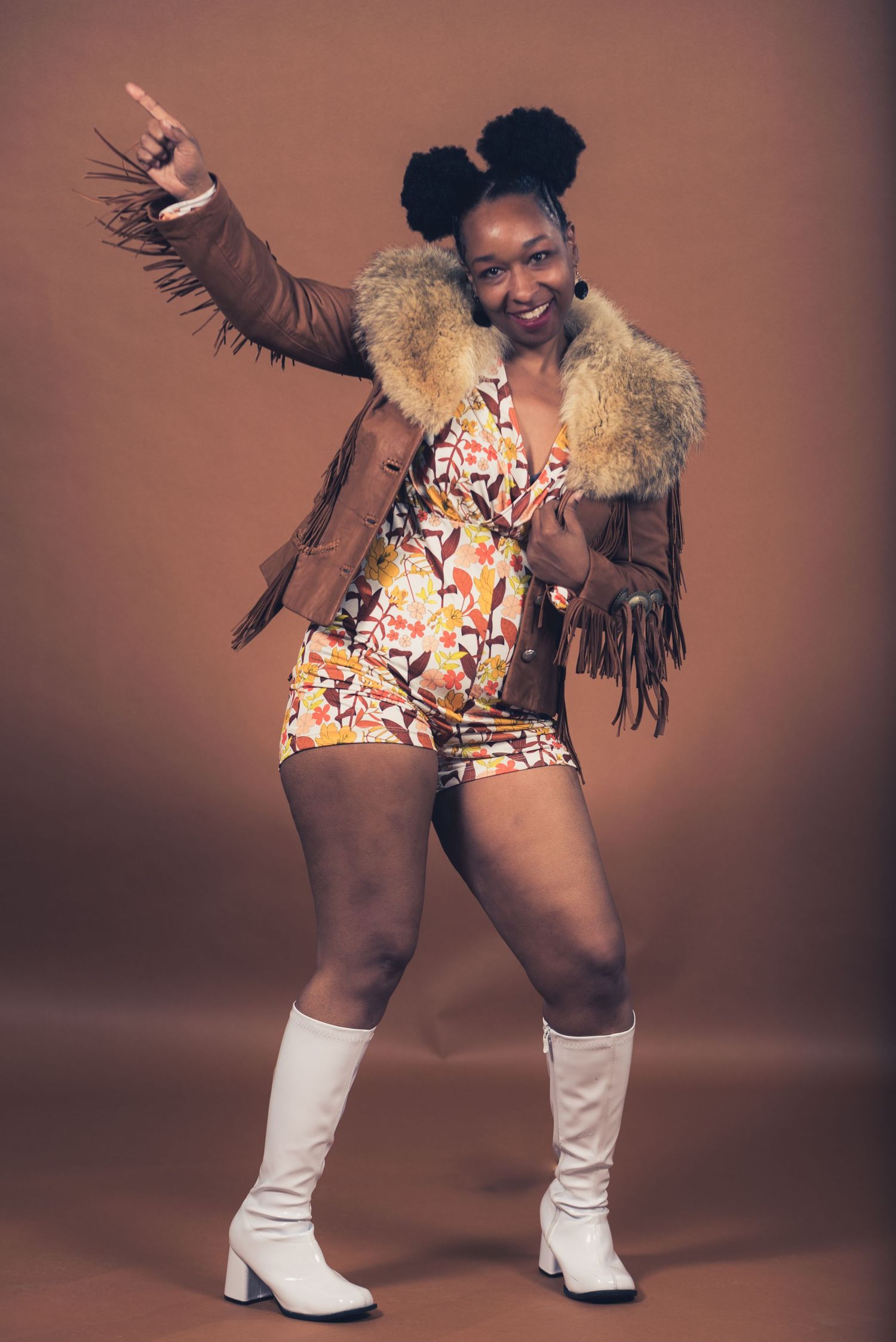 Energetic pose in brown fringe jacket, floral romper and white boots against studio backdrop.
