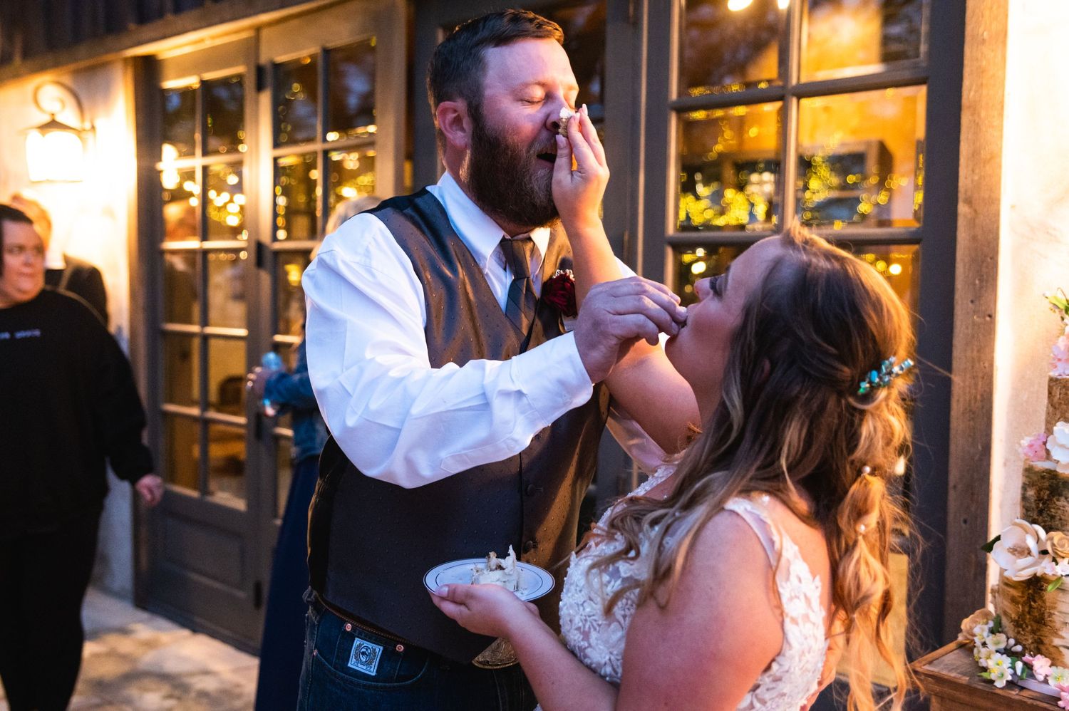 Wedding couple sharing cake in a playful moment at an evening reception venue.