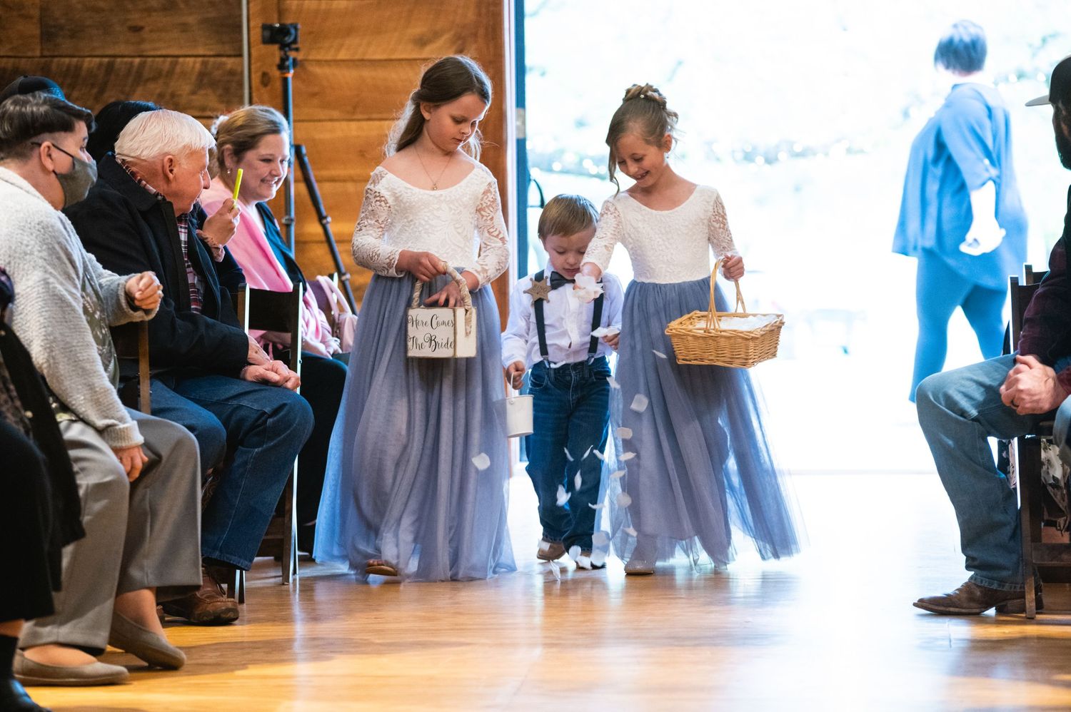 Young attendants walk down the aisle during a wedding ceremony in a rustic venue.