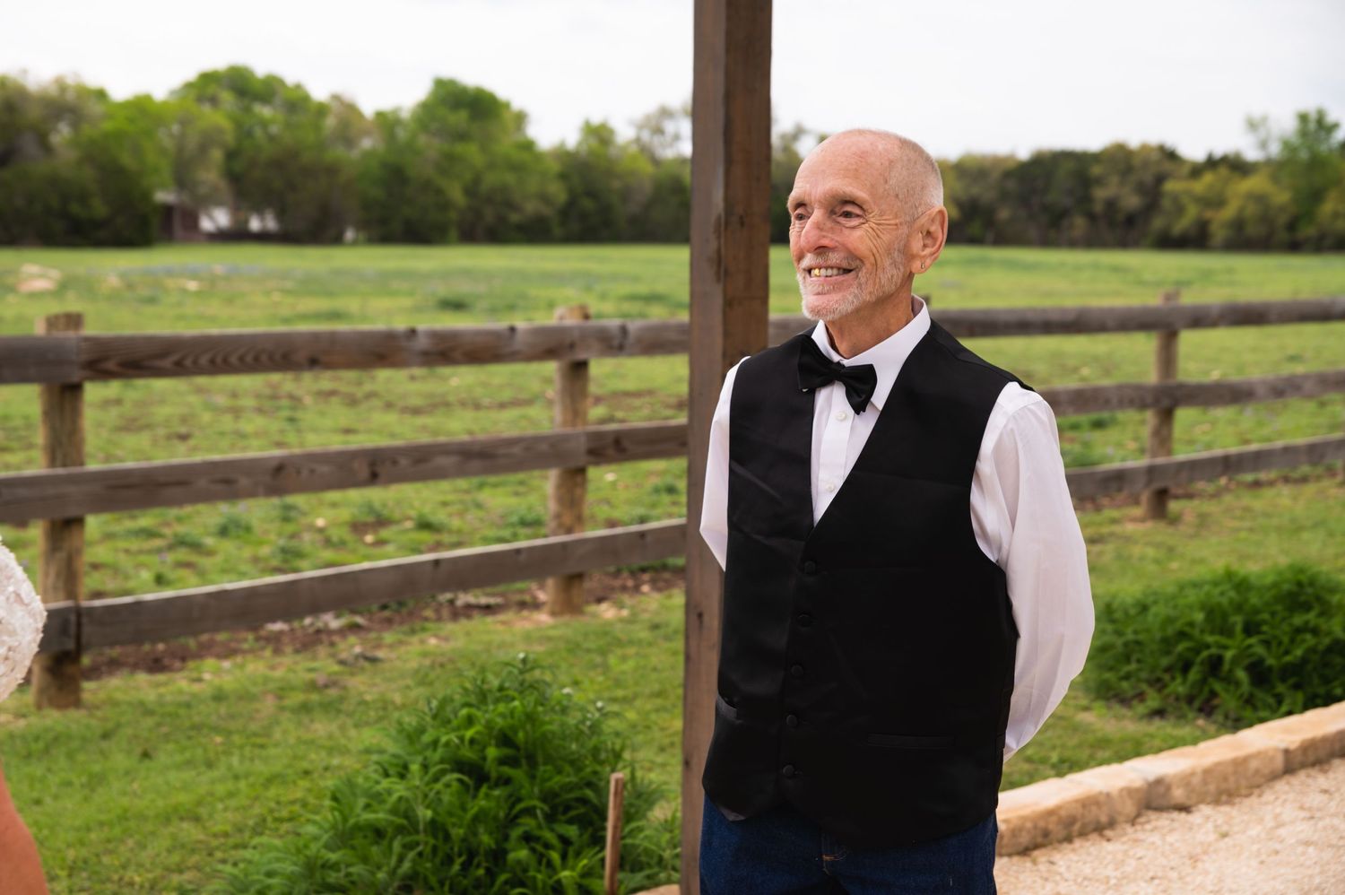 Gentleman in formal vest and bowtie smiling outdoors near rustic fencing.
