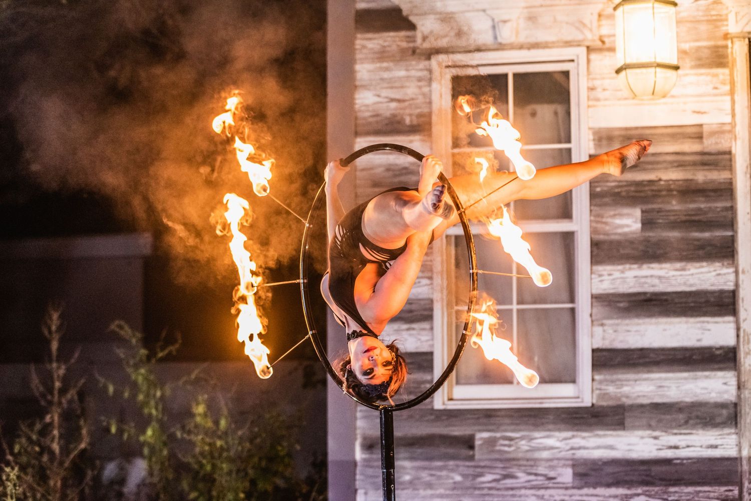 Circus performer executes dynamic pose on aerial hoop with fire elements against wooden structure.