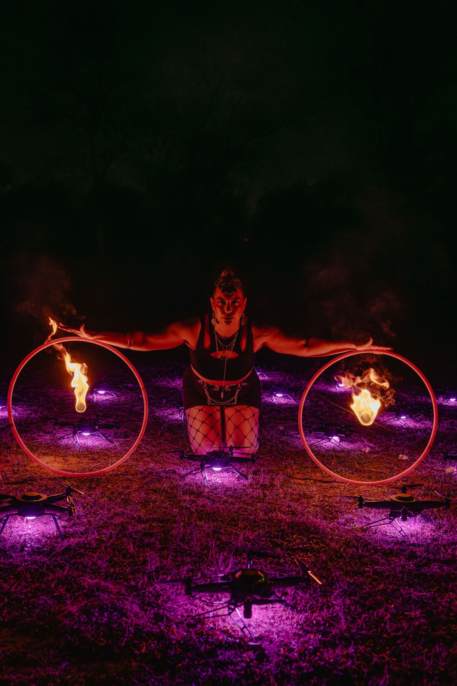Fire performer balances flaming hoops at night with purple lights illuminating the ground during an outdoor fire show.