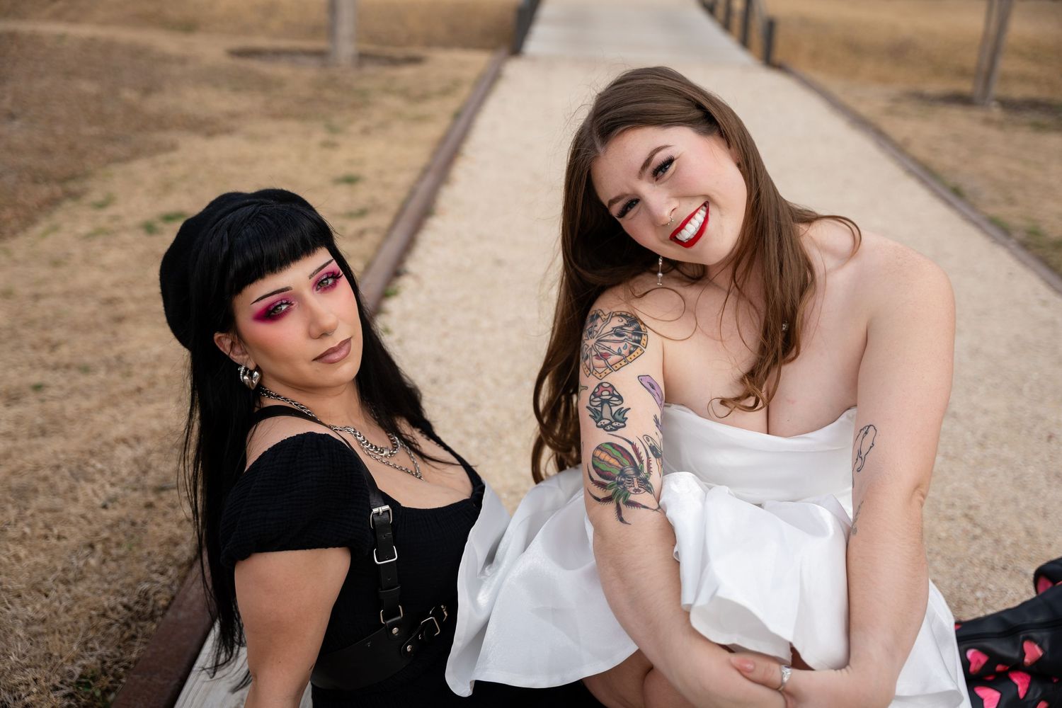 Two women smiling outdoors, one with bold makeup and black outfit, other in white dress with tattoos.