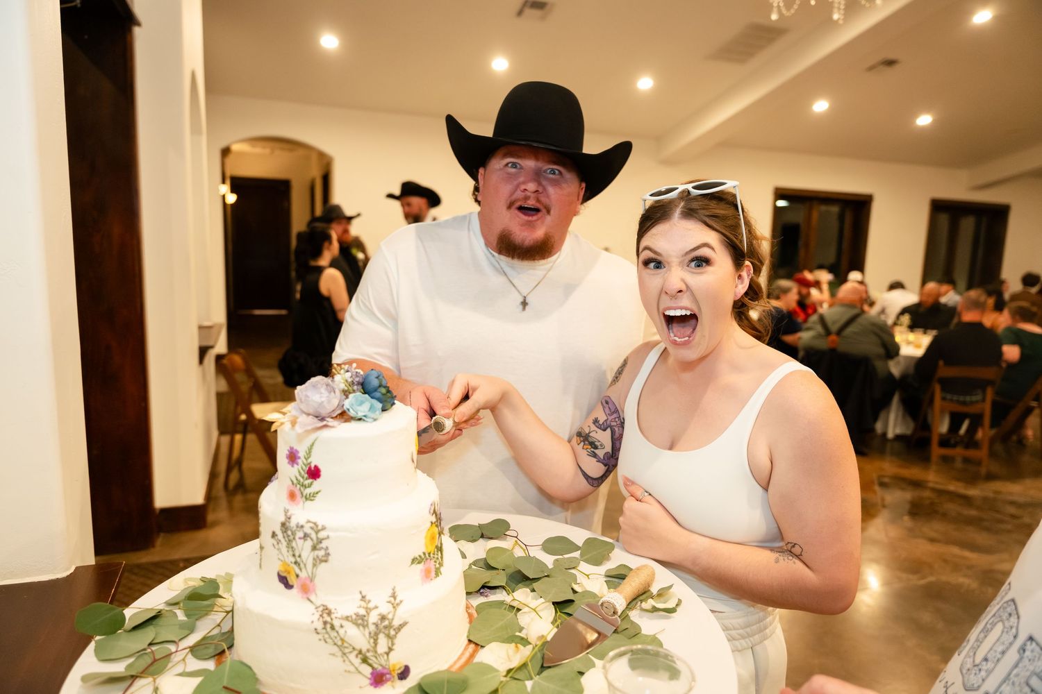 Bride and groom laughing while cutting white floral wedding cake indoors at elegant reception venue.