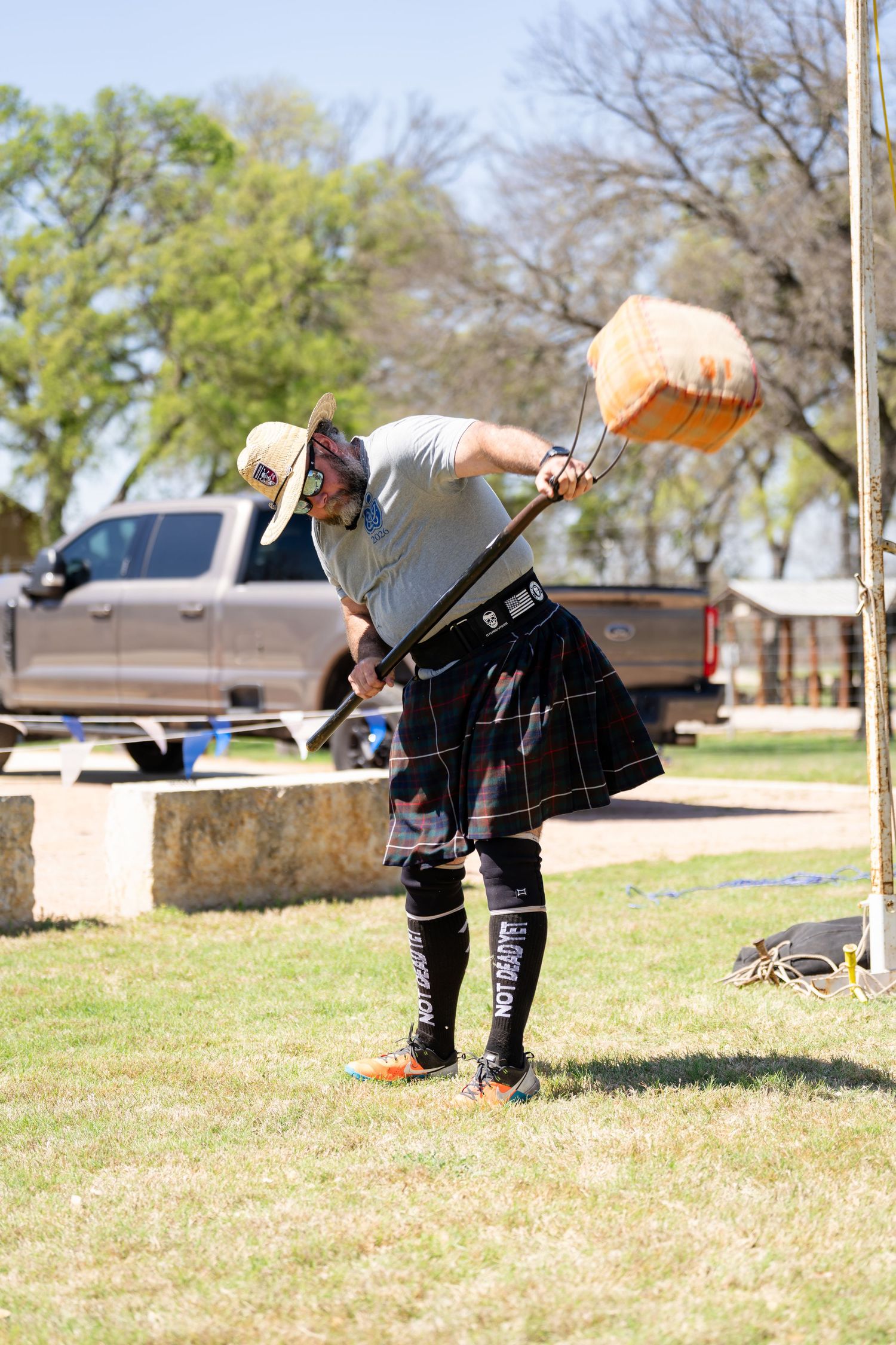 Man in tartan kilt and cap competes in Highland Games sheaf toss event outdoors.