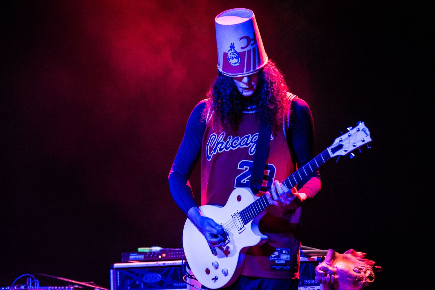 Guitarist performing on stage with a bucket on head in blue and red lighting atmosphere.