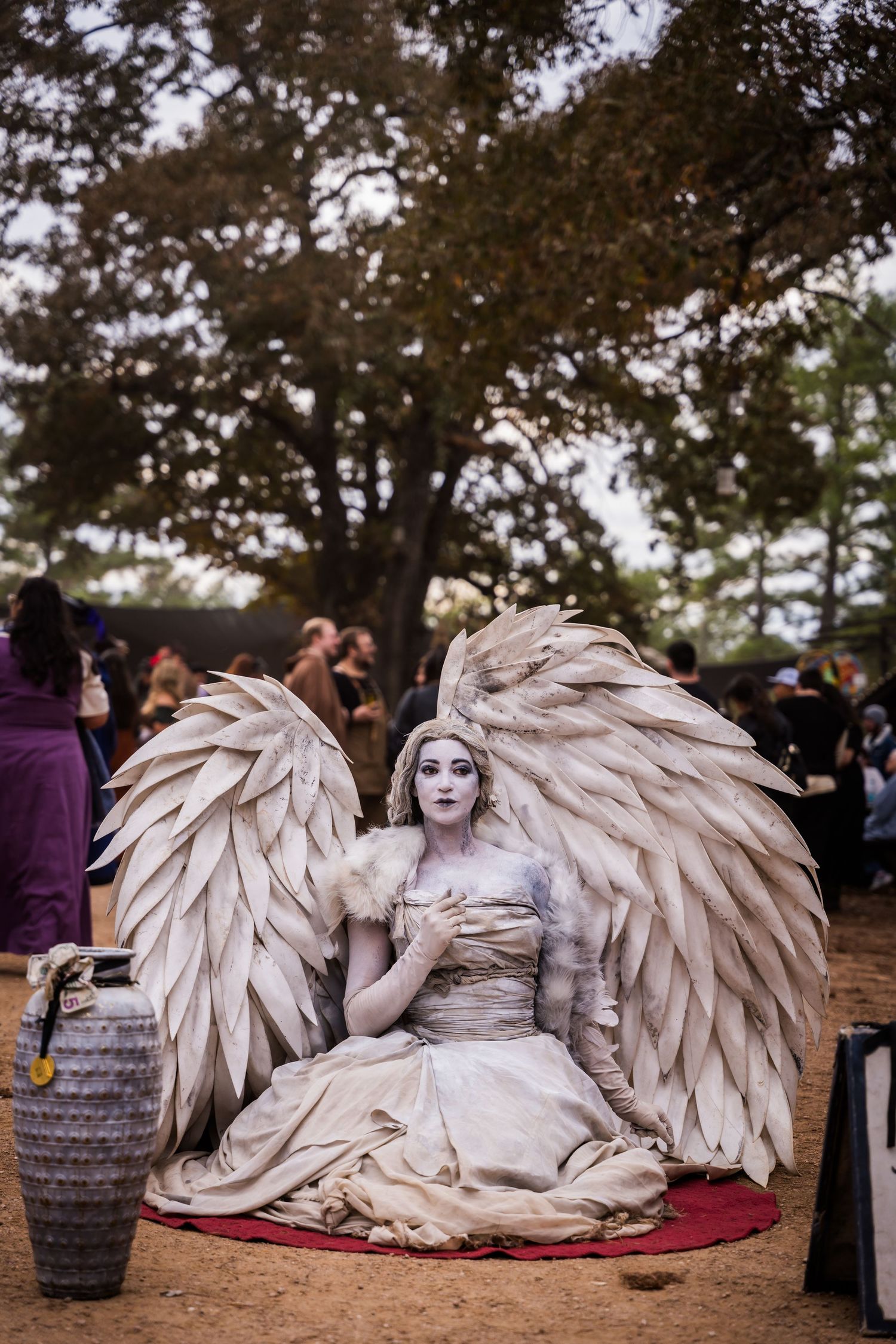 A performer in an ethereal white angel costume with large feathered wings rests on a red blanket outdoors.