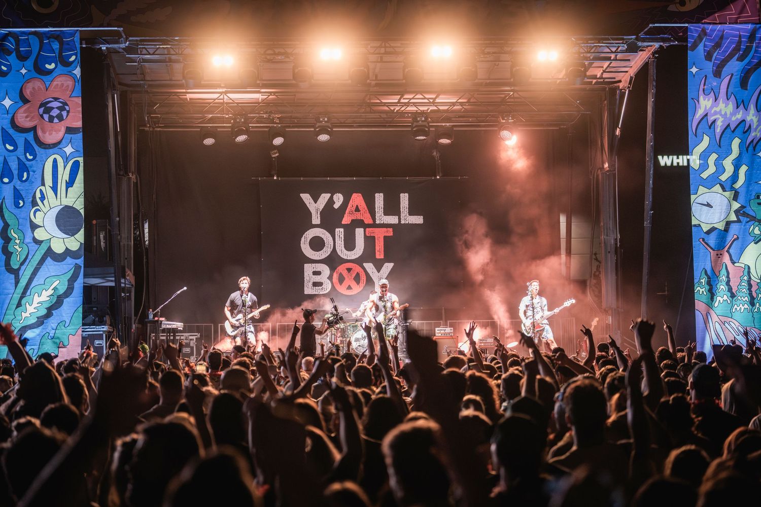 Concert crowd watches band perform on stage with 'Y'ALL OUT BOY' text display and dramatic lighting effects.