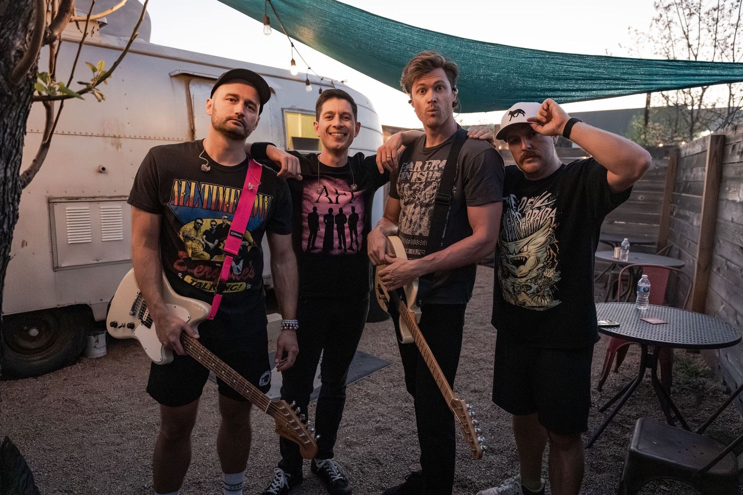 Four young men in band t-shirts pose together outdoors near a vintage trailer at a music event.