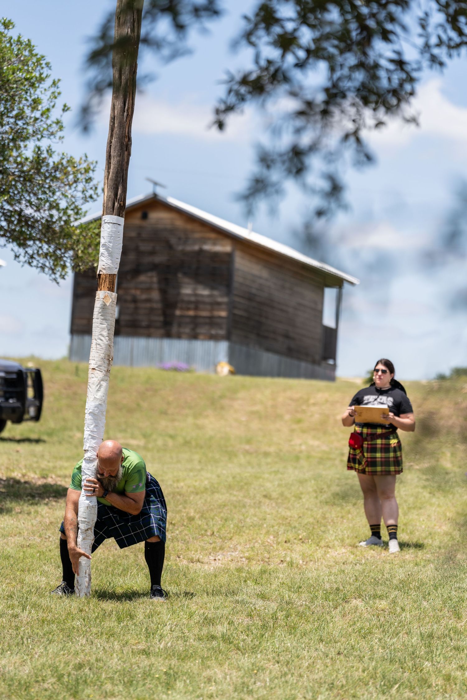 Man in kilt tossing caber at Scottish Highland Games with barn and judge in tartan skirt in background.