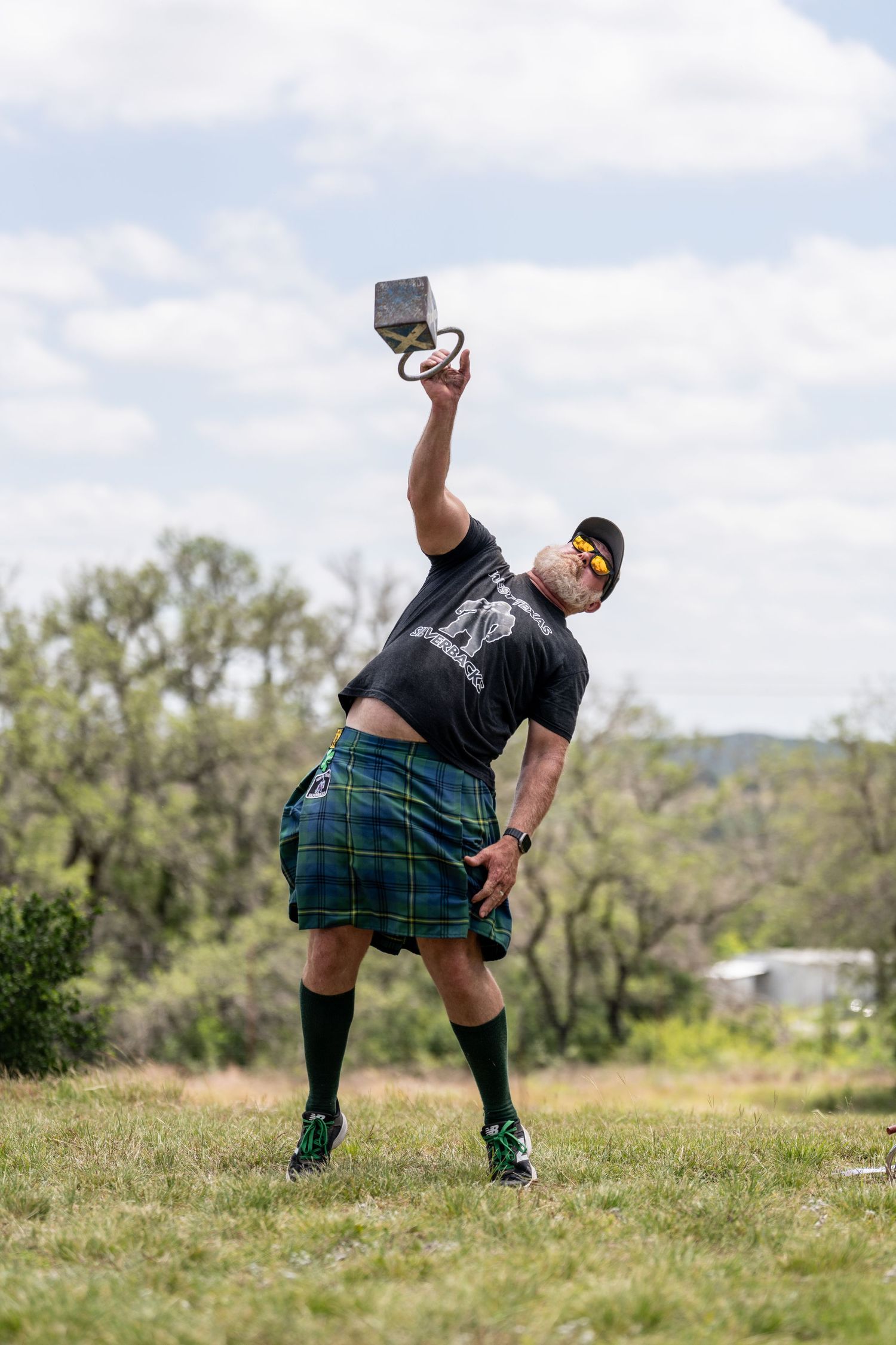 Man in tartan kilt performing Highland Games weight throw outdoors, wearing sunglasses and black shirt on green field.