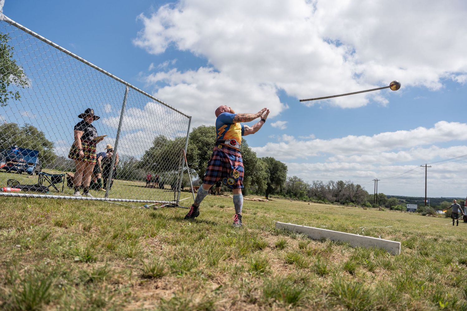 Athlete in tartan kilt throws hammer at Highland Games competition, with chain-link fence and spectators in background.