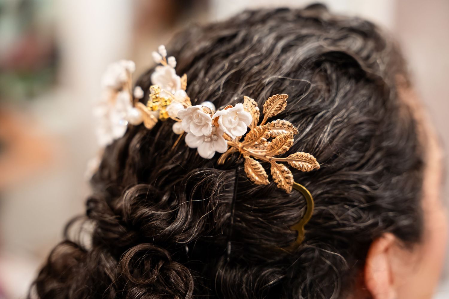 Close-up of a bridal hair accessory featuring white flowers and gold leaves on dark curly hair.