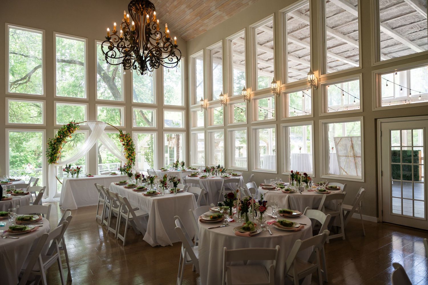 Elegant wedding reception hall with white round tables, floral arch, chandelier, and floor-to-ceiling windows.