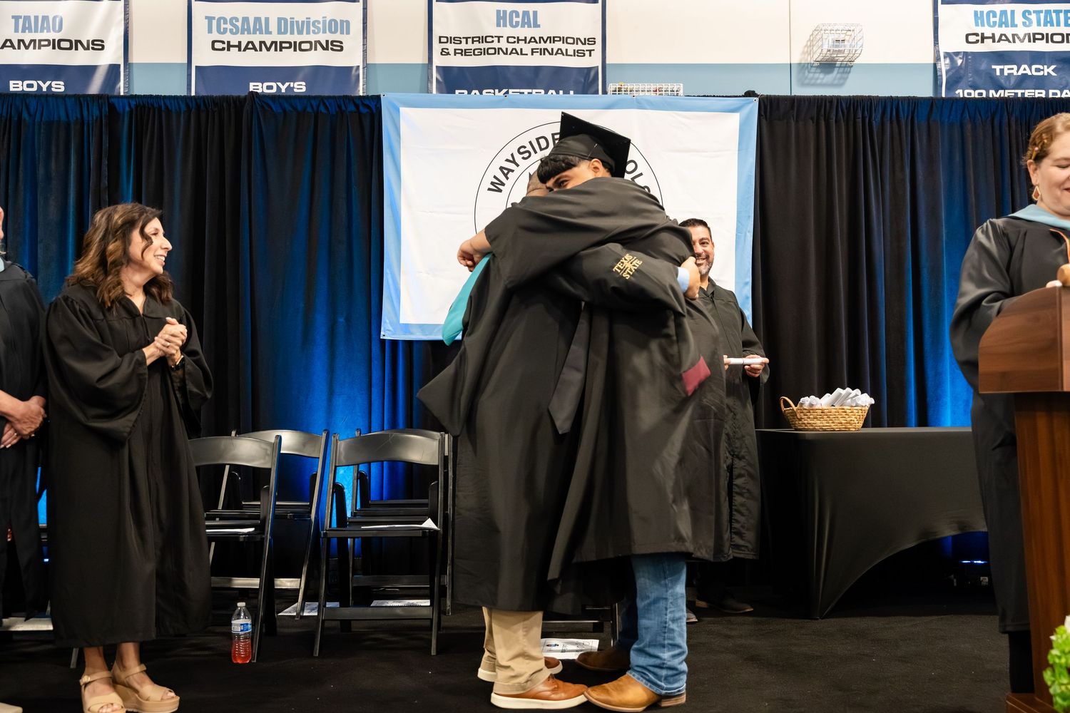 Two graduates in caps and gowns share a warm hug on stage during a graduation ceremony indoors.