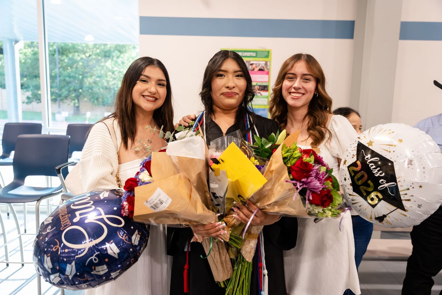 Three smiling graduates in white gowns holding colorful flower bouquets and balloons at a graduation celebration.