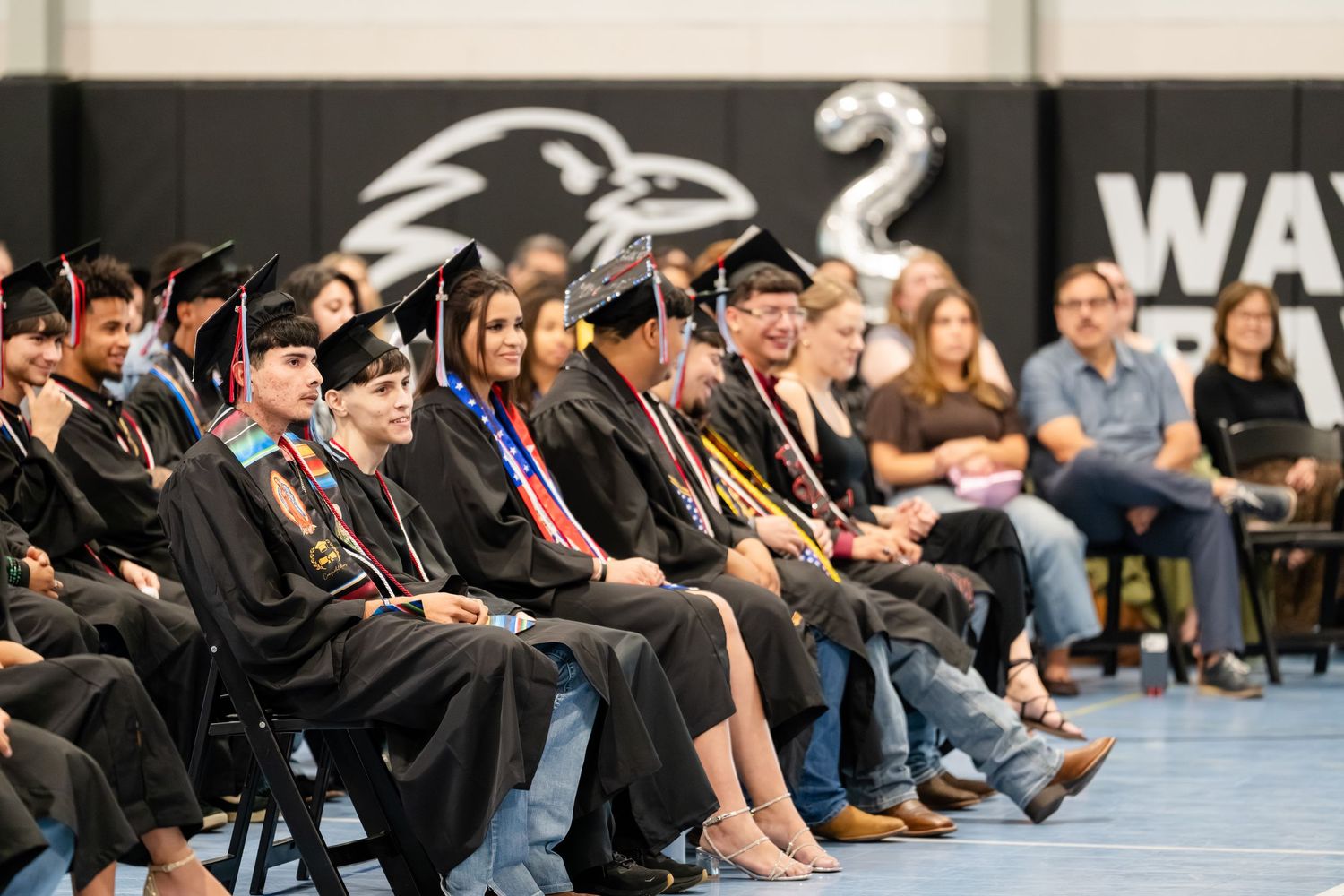 College graduates in black caps and gowns seated at an indoor graduation ceremony in a school gymnasium.