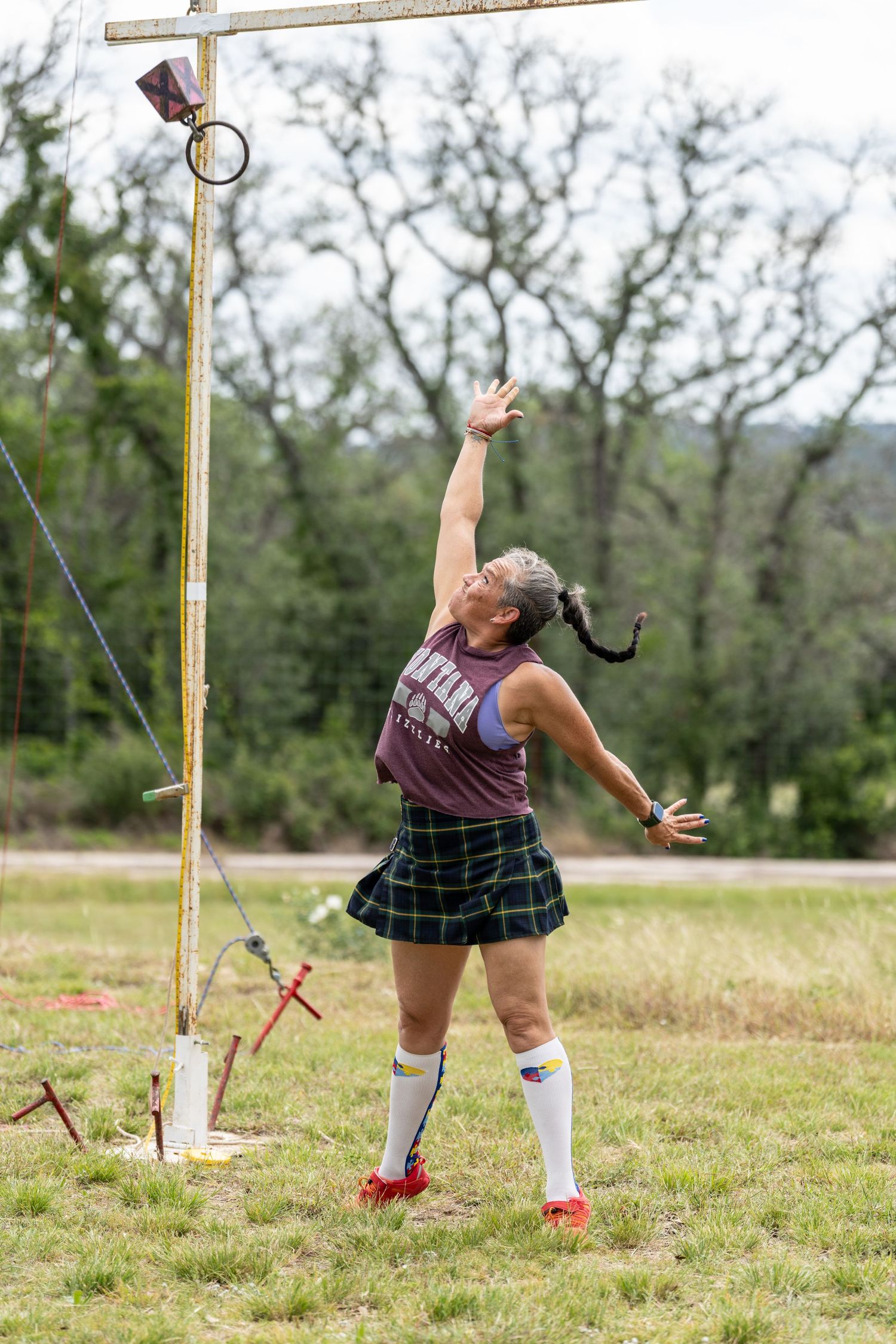 Woman in tartan kilt and white socks reaches up during Highland Games competition outdoors.