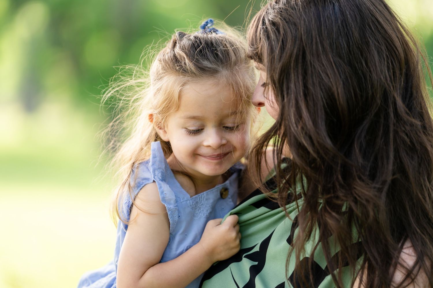 A mother tenderly kisses her smiling young daughter in a sunny green park, both wearing blue and green outfits.