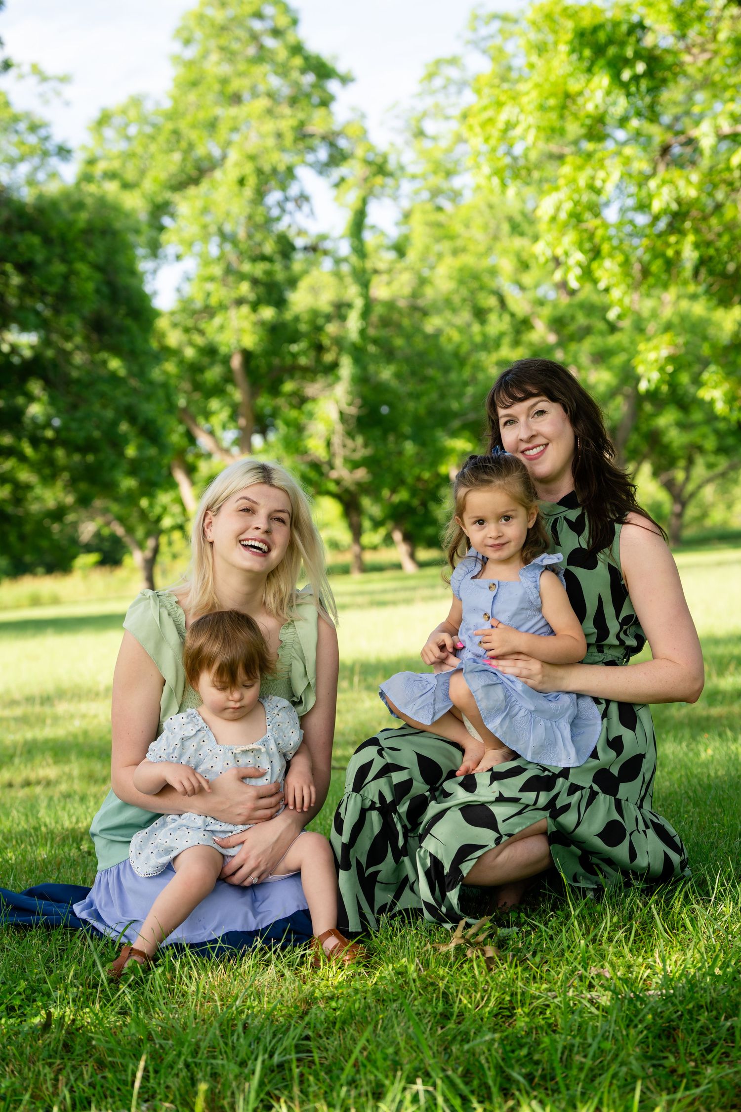 Two mothers sitting on green grass in a park, each holding a young daughter, smiling on a sunny day.