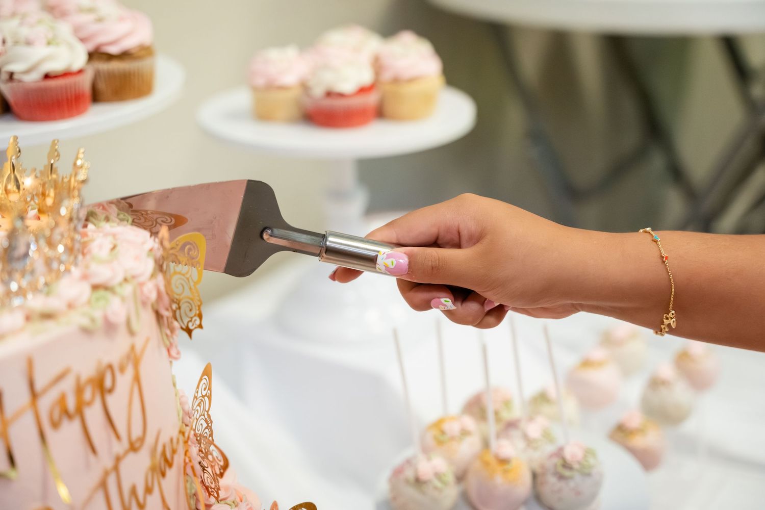 Hand cutting a pink birthday cake with a spatula near cake pops and cupcakes on a dessert table.