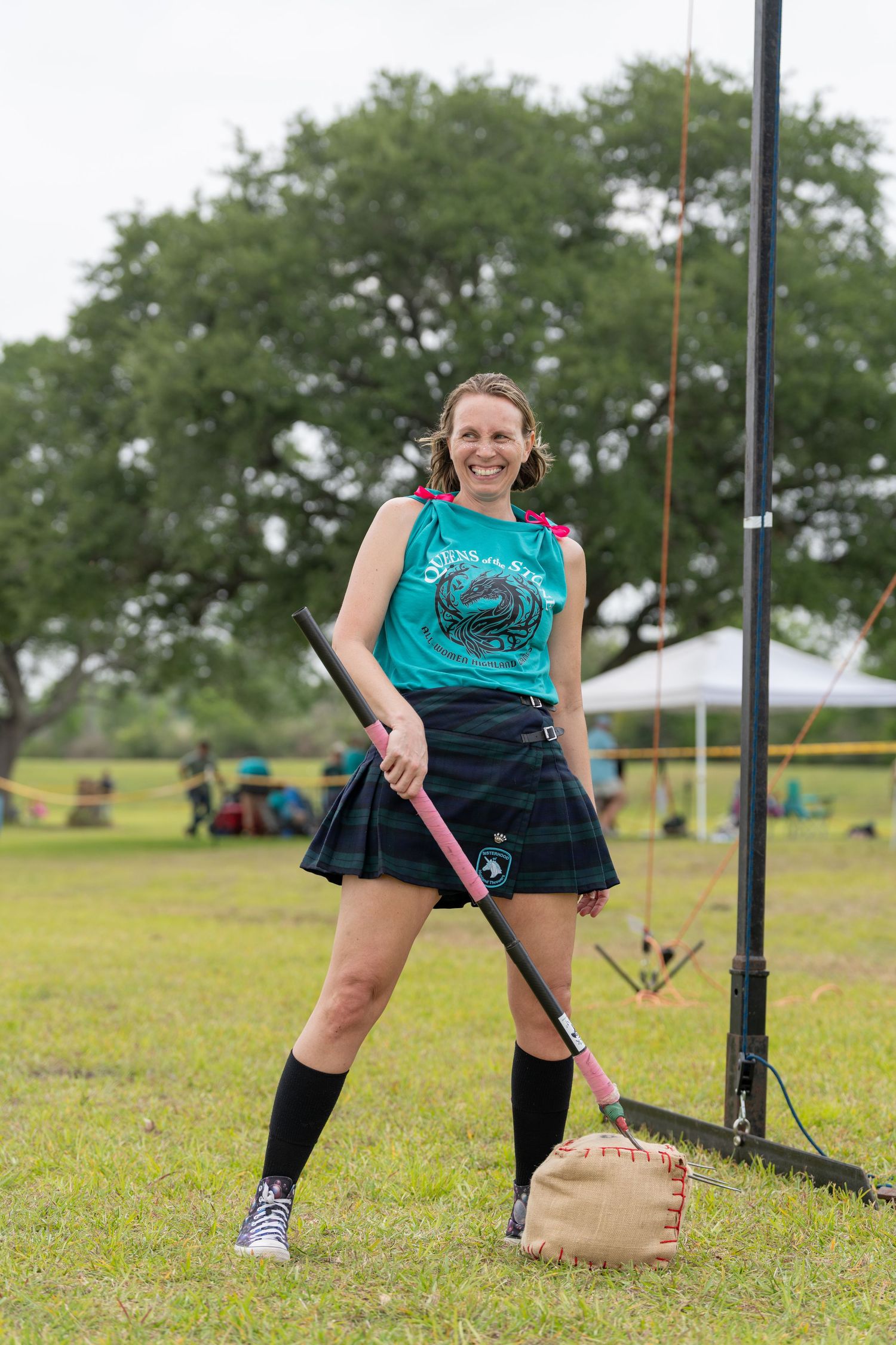 Smiling woman in tartan kilt and teal top holds a pink hammer at an outdoor Highland Games competition.