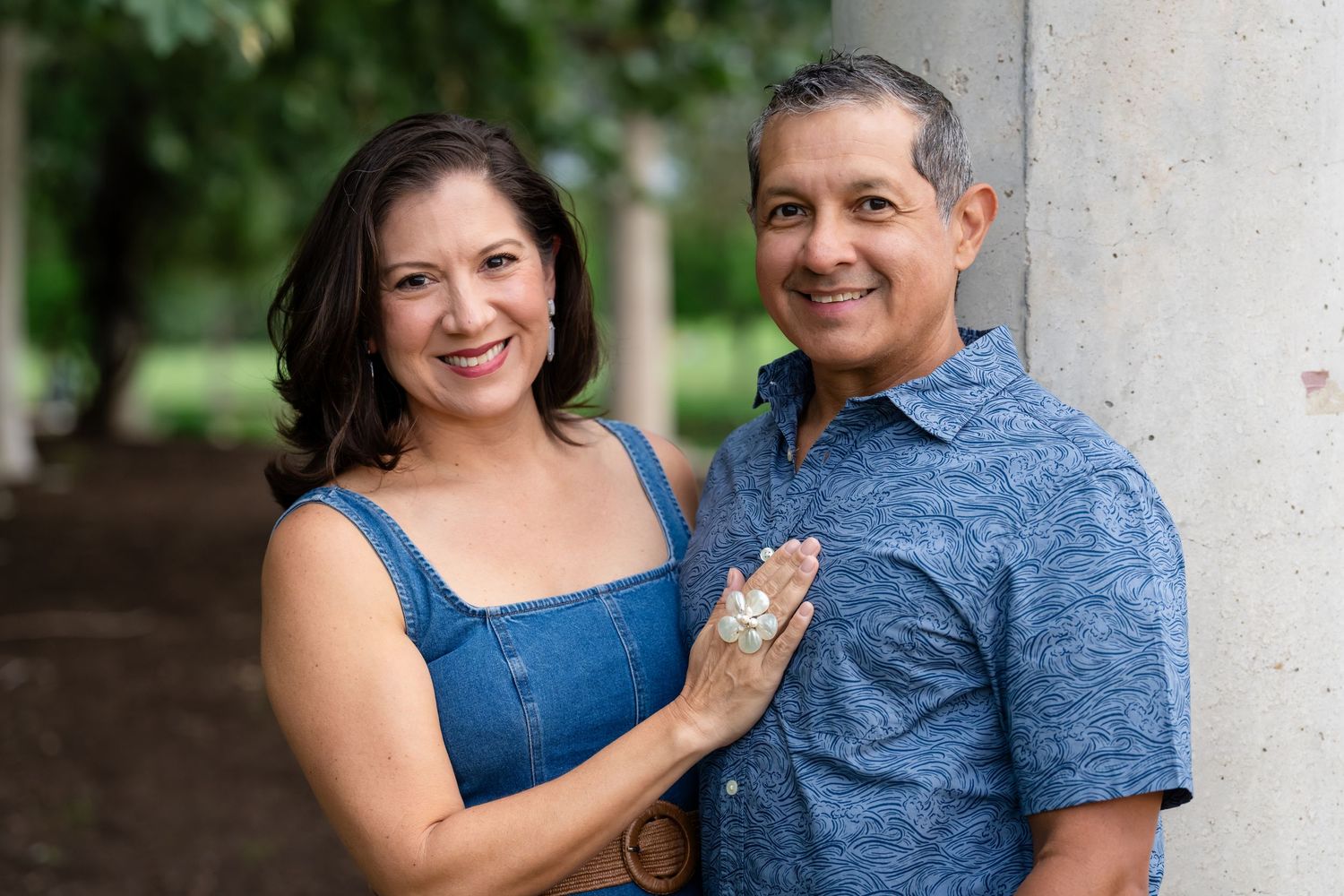 A smiling Hispanic couple poses outdoors, woman in denim top and man in blue floral shirt, leaning against a column.