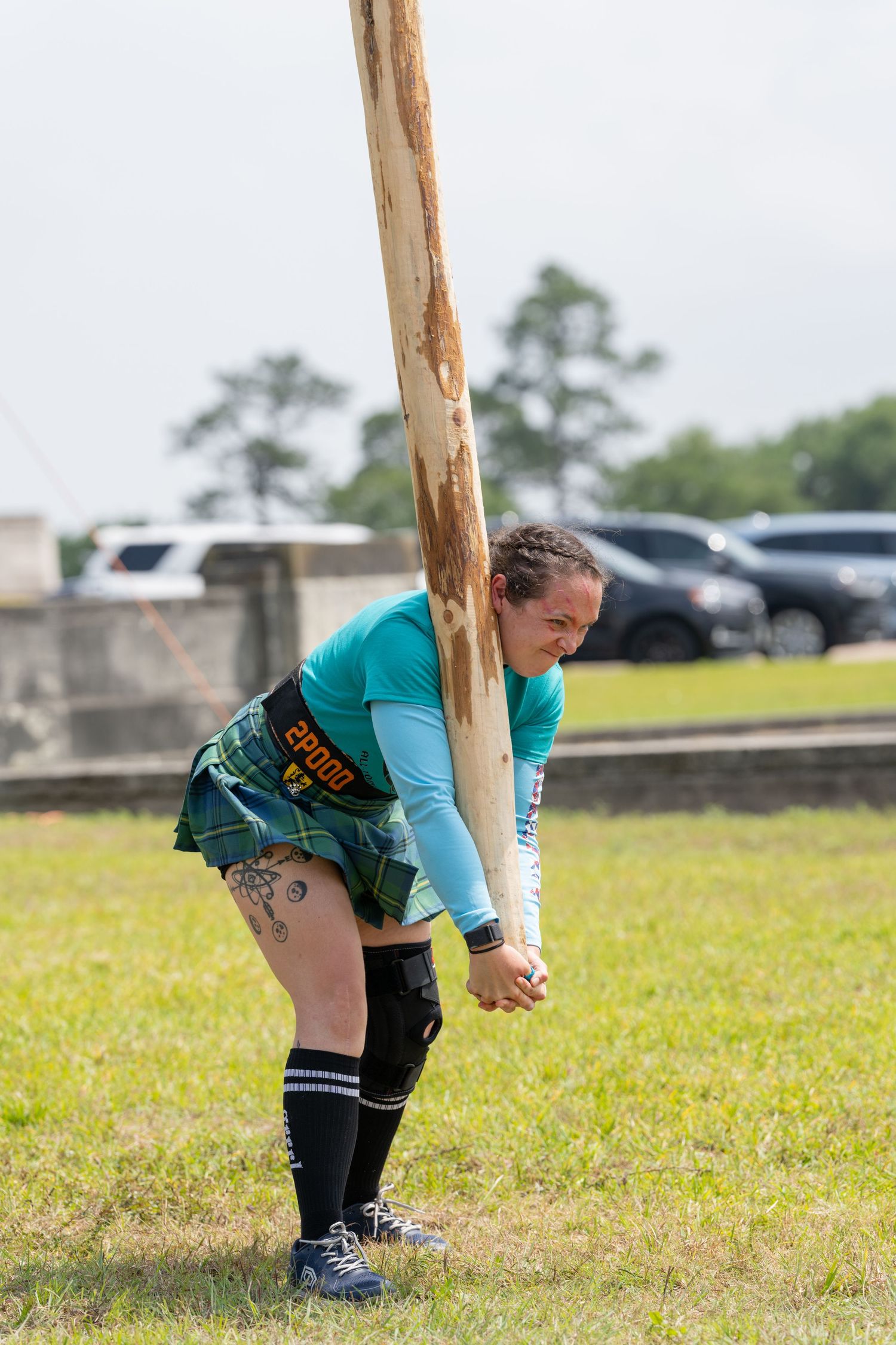 Woman in teal shirt and kilt competing in caber toss at Highland Games event outdoors.