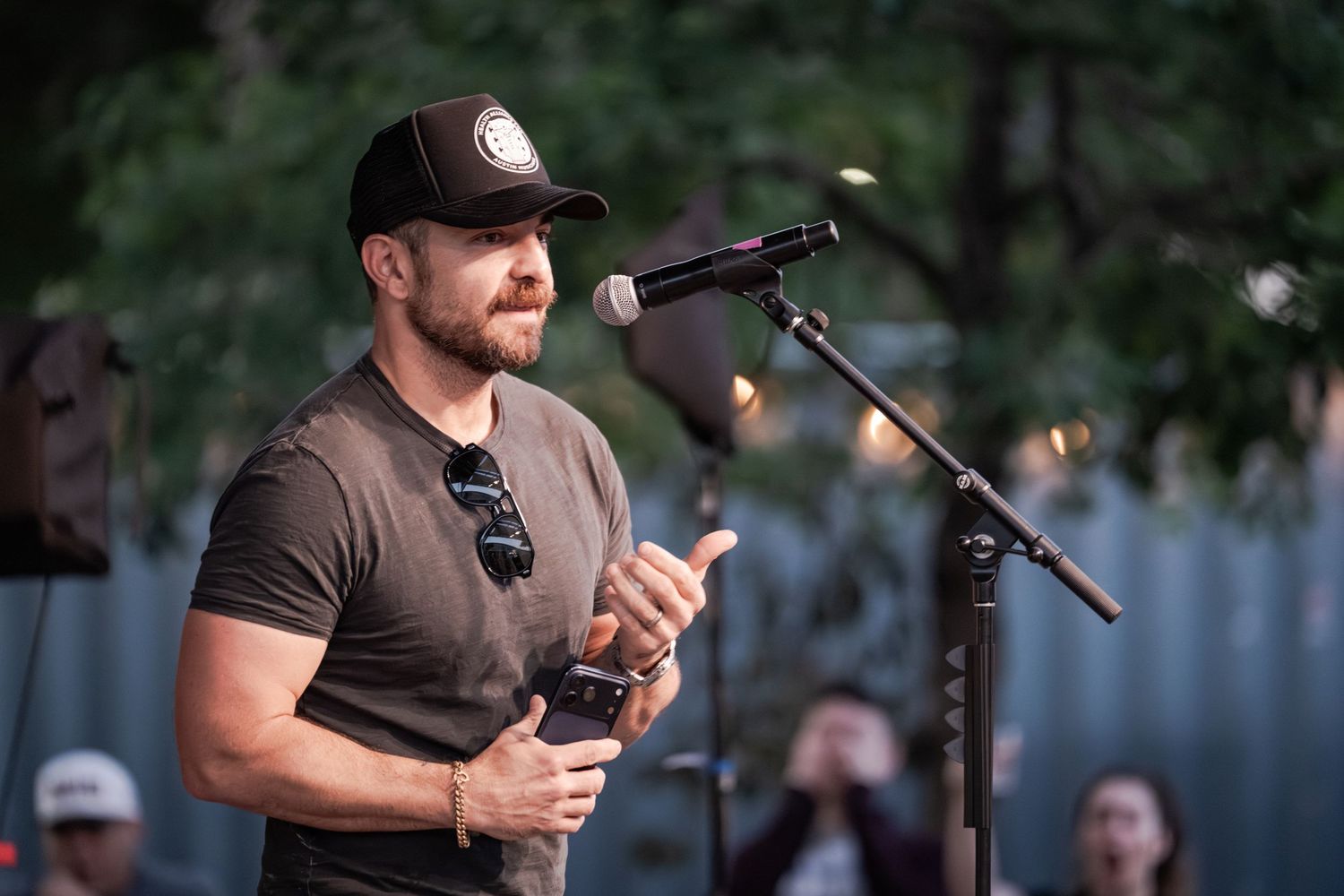 Man in black cap and t-shirt speaking at outdoor microphone stand during evening event.