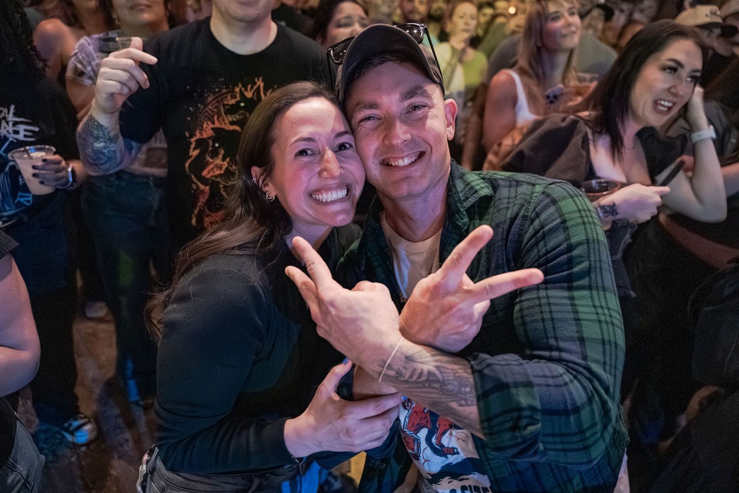 Two friends smile and flash rock-on hand signs at a lively concert event.