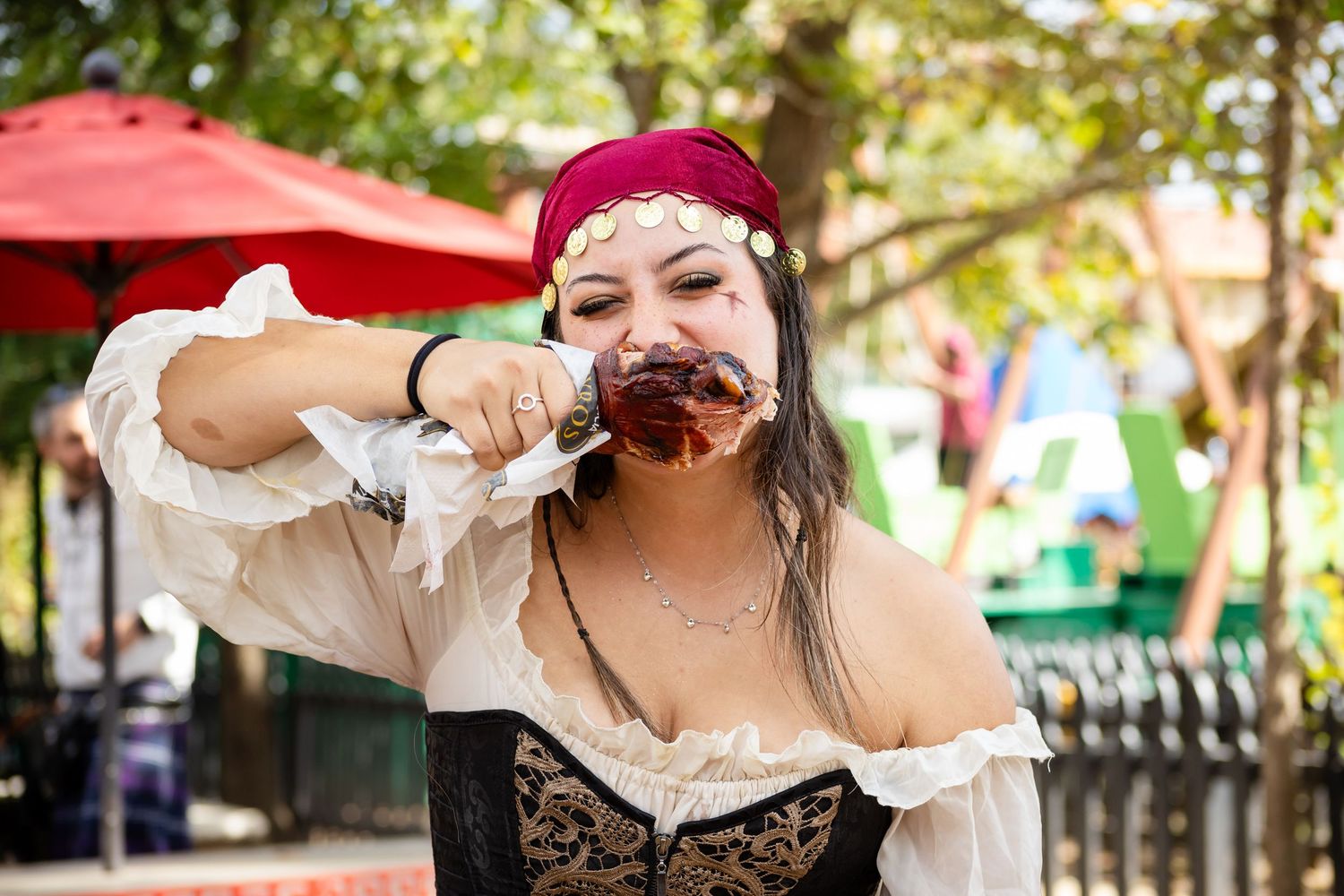 A performer in pirate costume with a red bandana takes a theatrical bite of food at an outdoor renaissance festival.