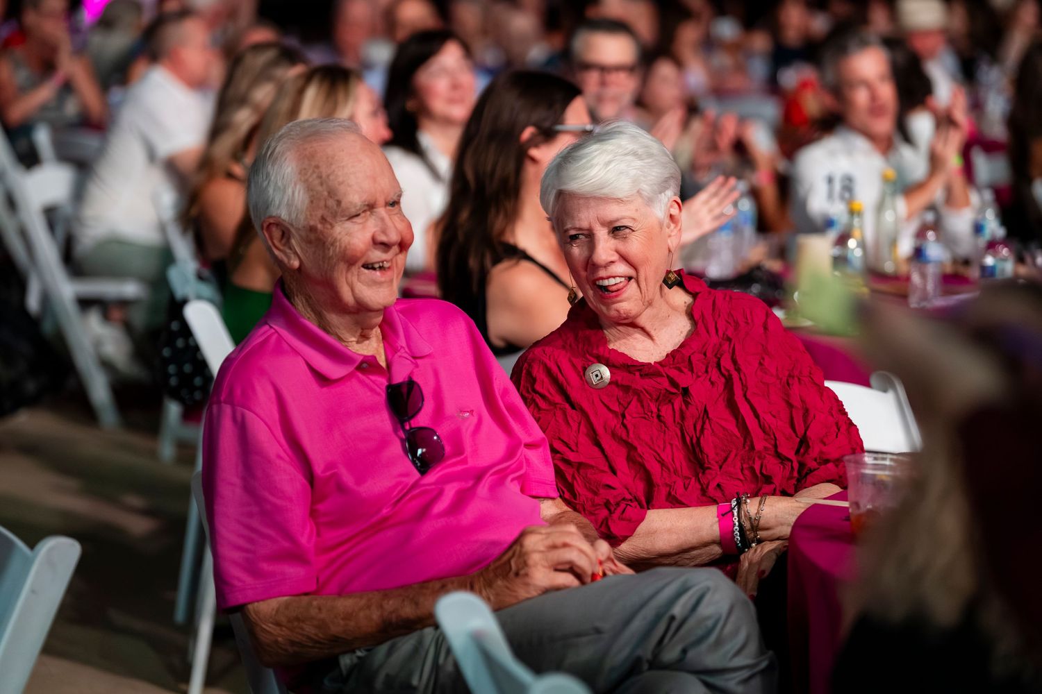 Elderly couple wearing matching bright pink sweaters share a tender moment of laughter together at a social gathering.