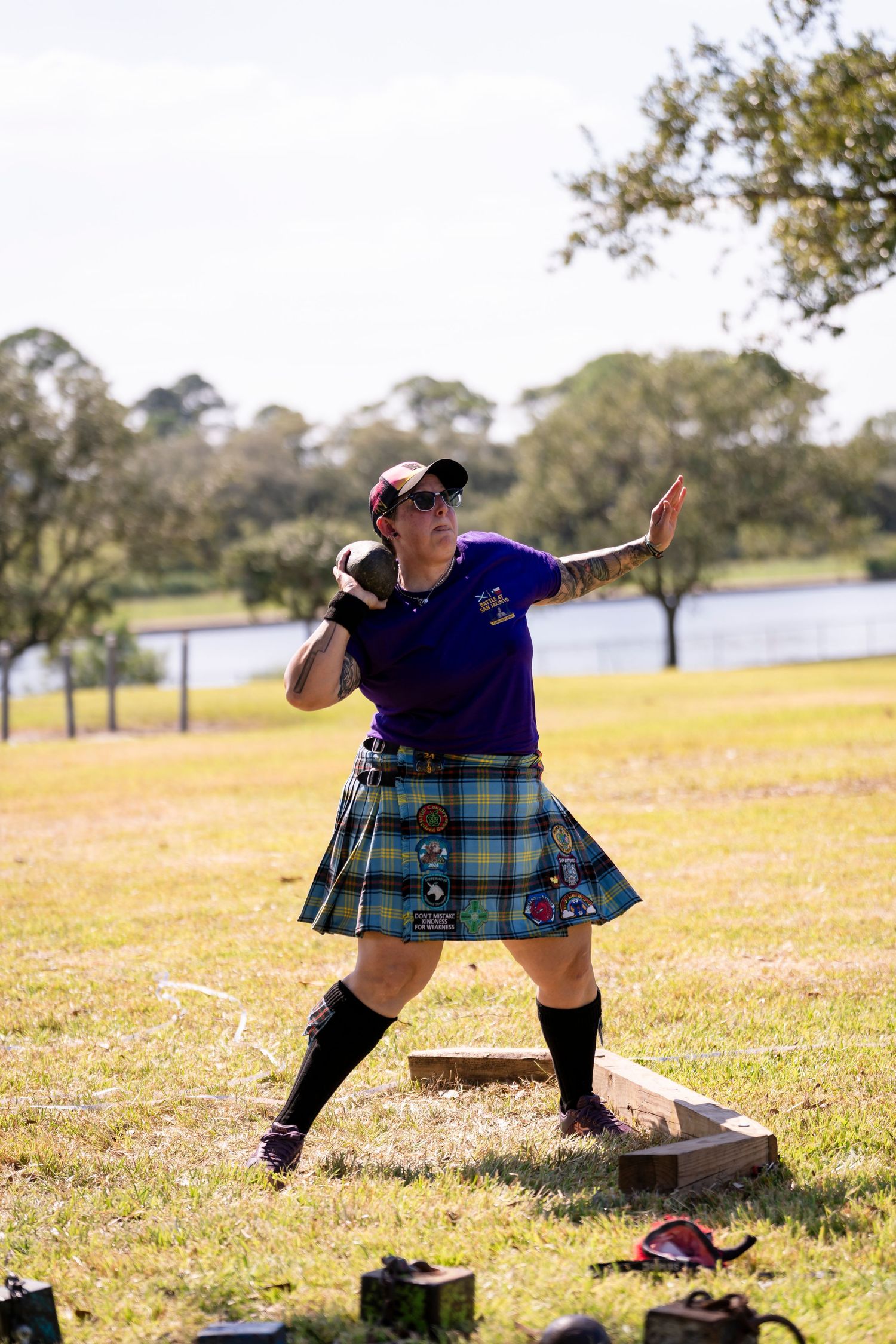 A person in a plaid kilt performs traditional Scottish dance moves in a grassy outdoor field on a sunny day.
