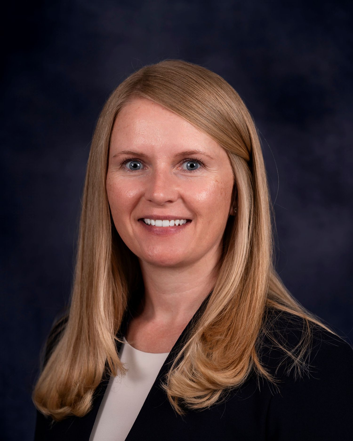 Professional headshot of someone with blonde hair and a warm smile against a dark background.