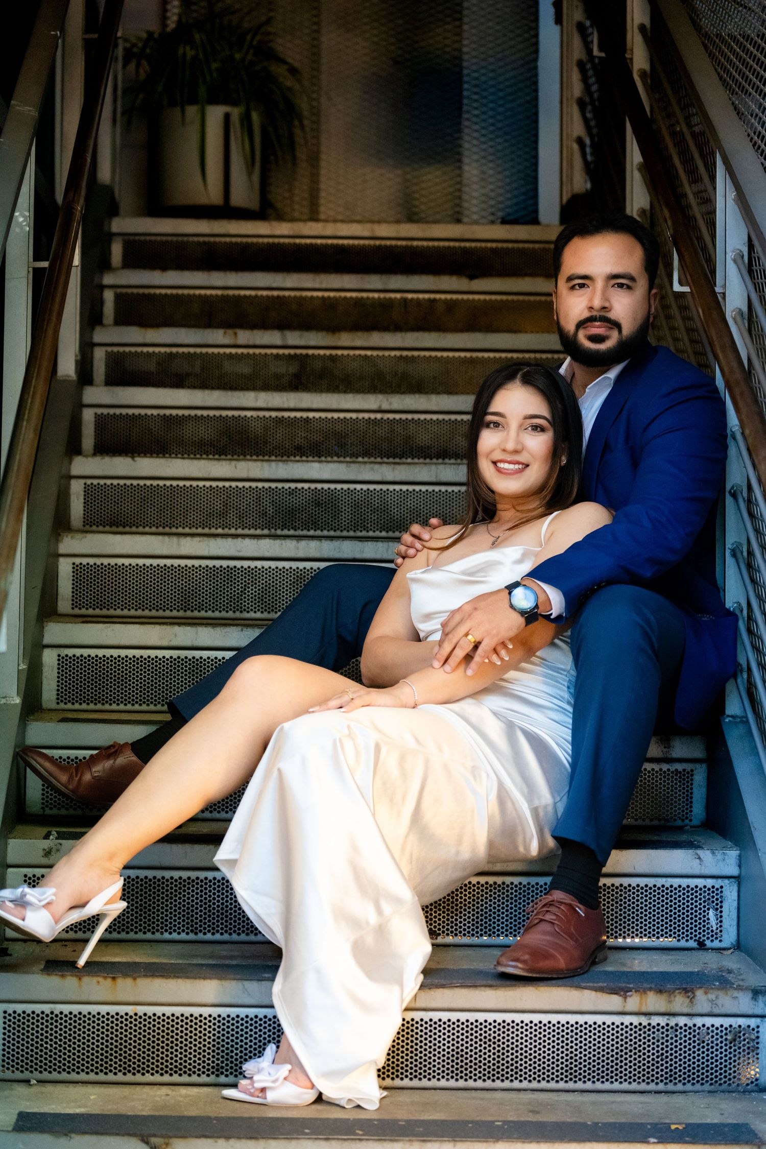 A couple relaxes on industrial metal stairs, with the person wearing a white dress leaning against their partner in blue.