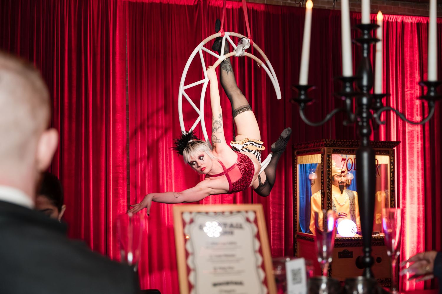 A circus performer gracefully hangs from a geometric aerial hoop against a red curtain backdrop with lanterns.