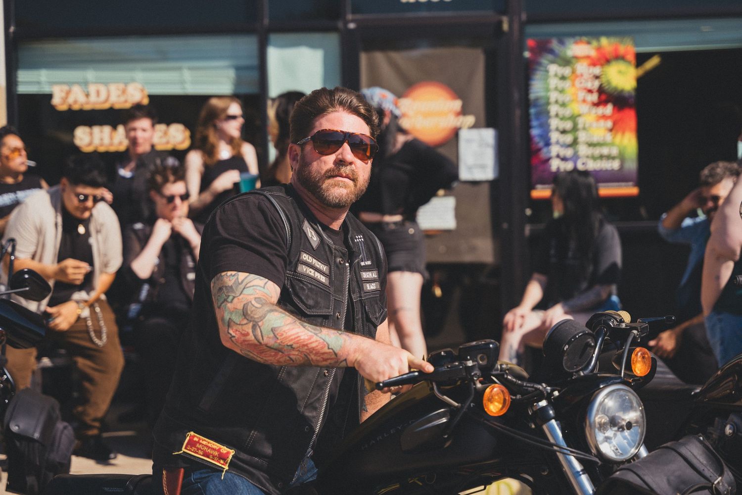 Bikers and motorcycle enthusiasts gather at a crowded outdoor event with bikes and vendors visible in the background.