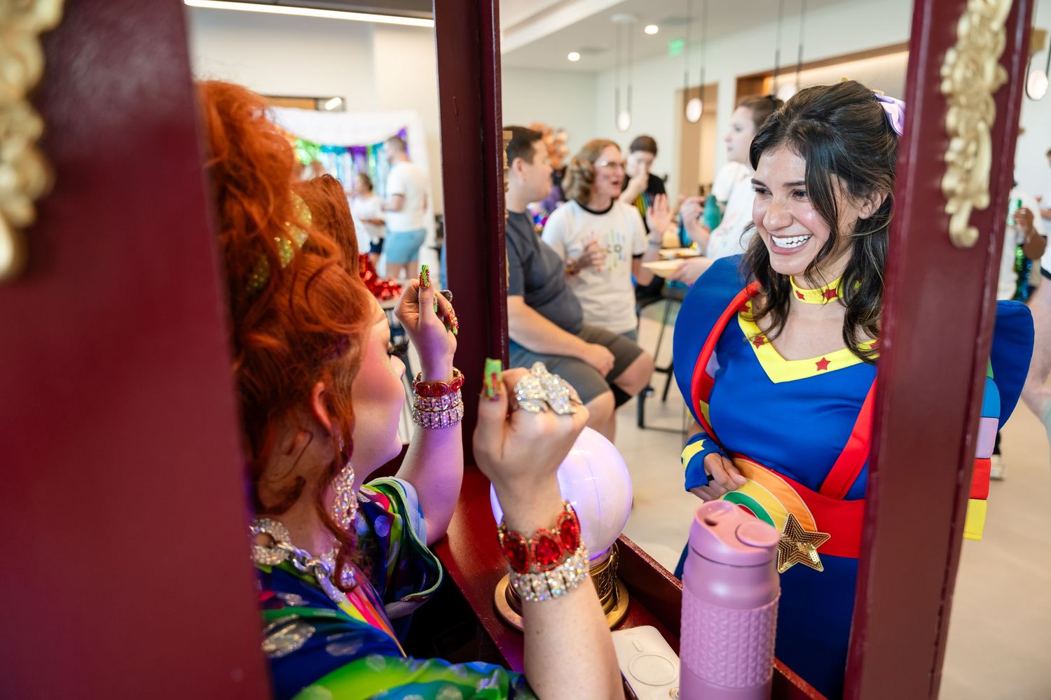 Two women in colorful costumes smile at each other during a lively cosplay or themed event indoors.