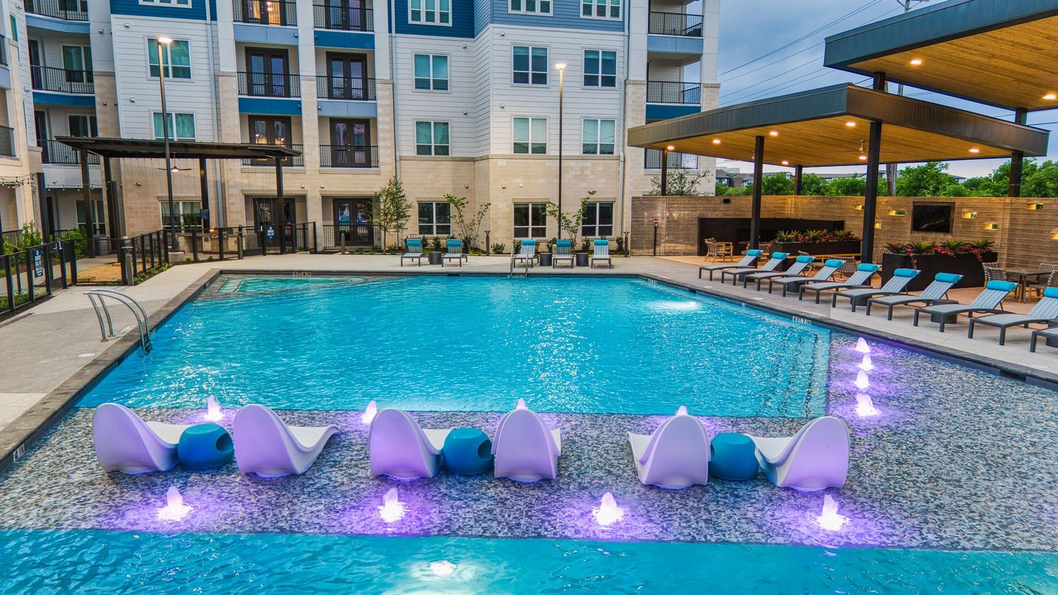 Resort-style swimming pool with purple LED lighting and in-water loungers at apartment complex.