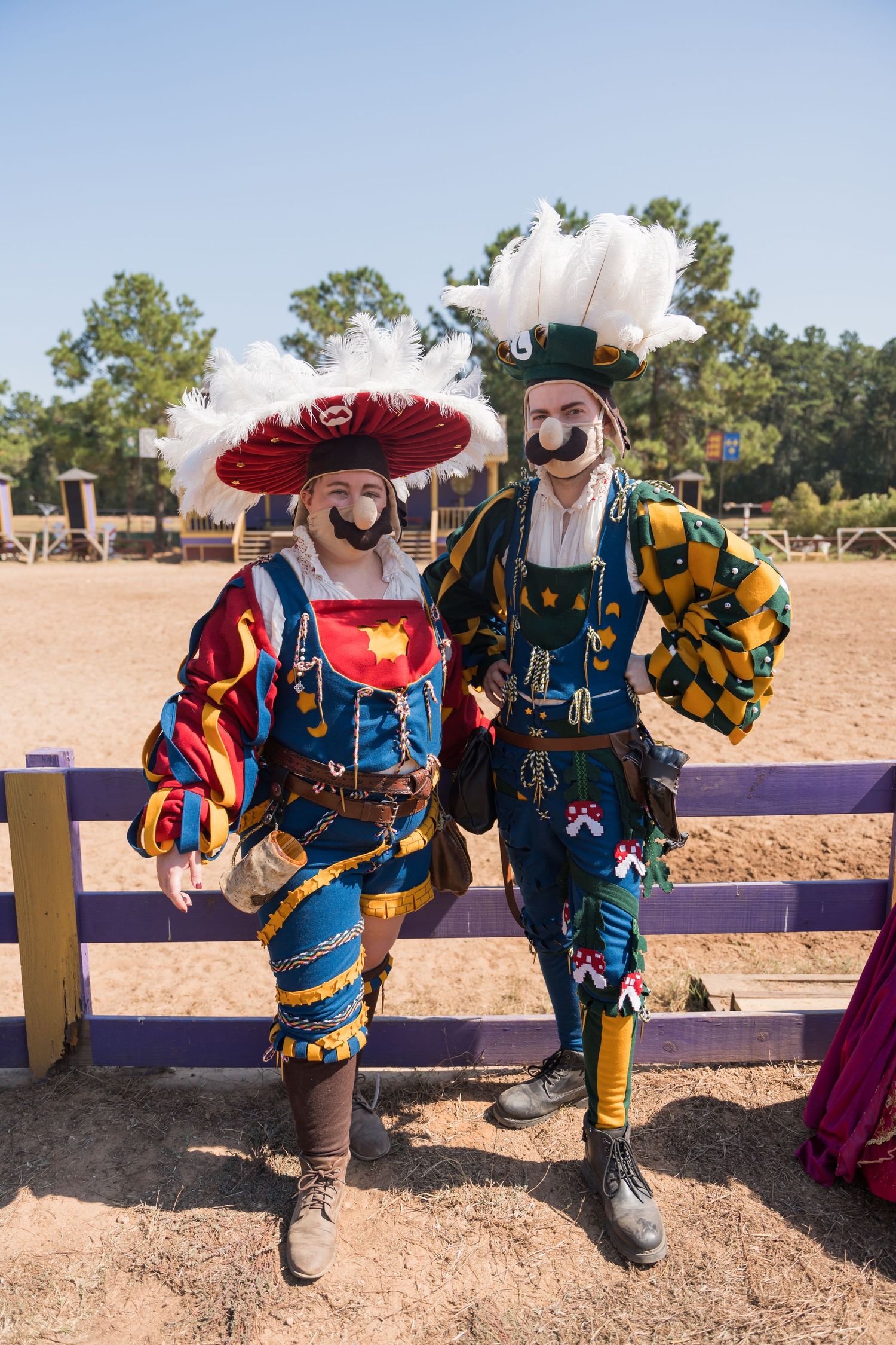Two performers in colorful renaissance costumes with elaborate plumed hats pose together at a fair.