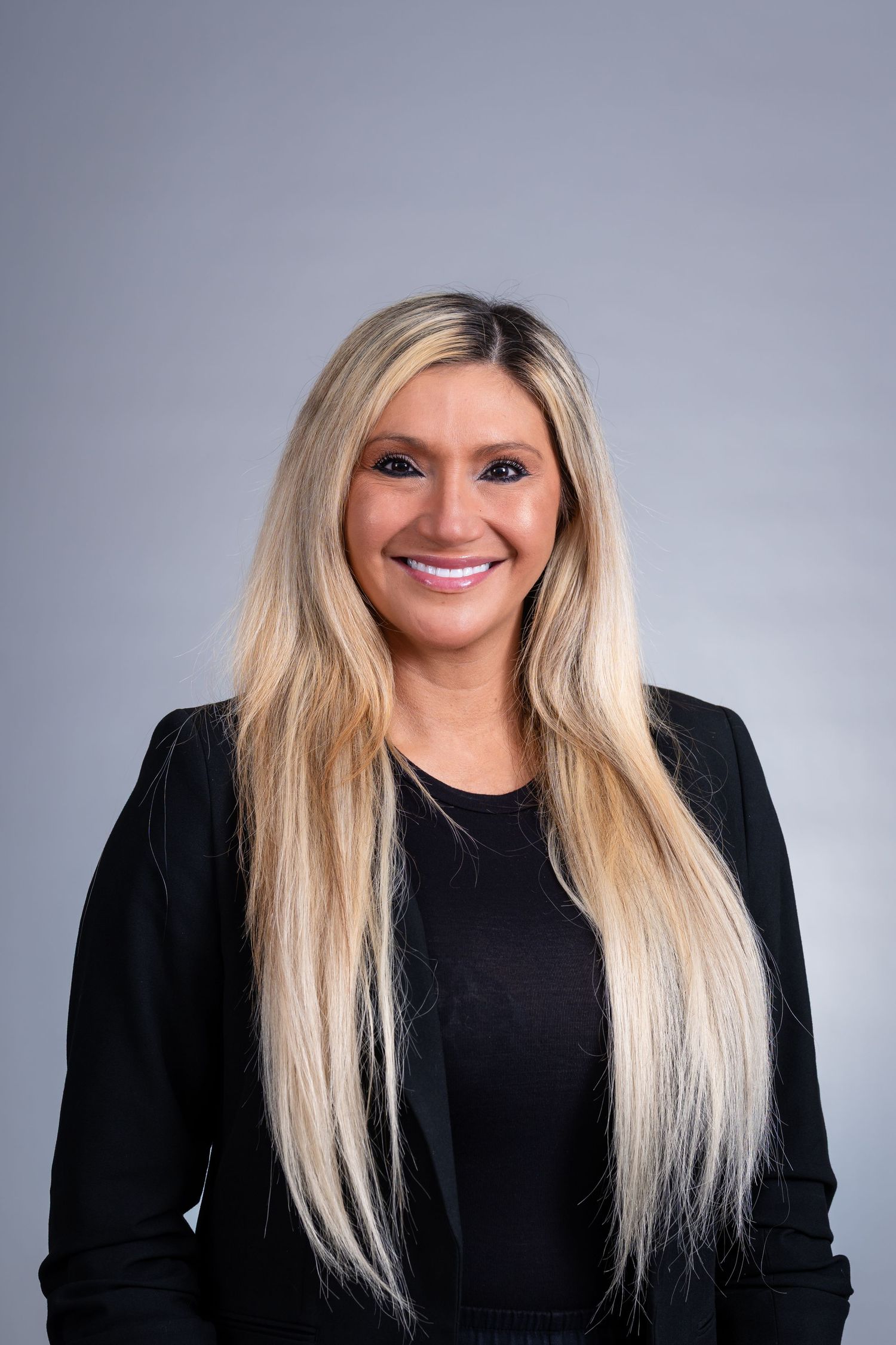 Professional headshot of a person with long blonde hair wearing a black blazer against a gray background, smiling warmly.