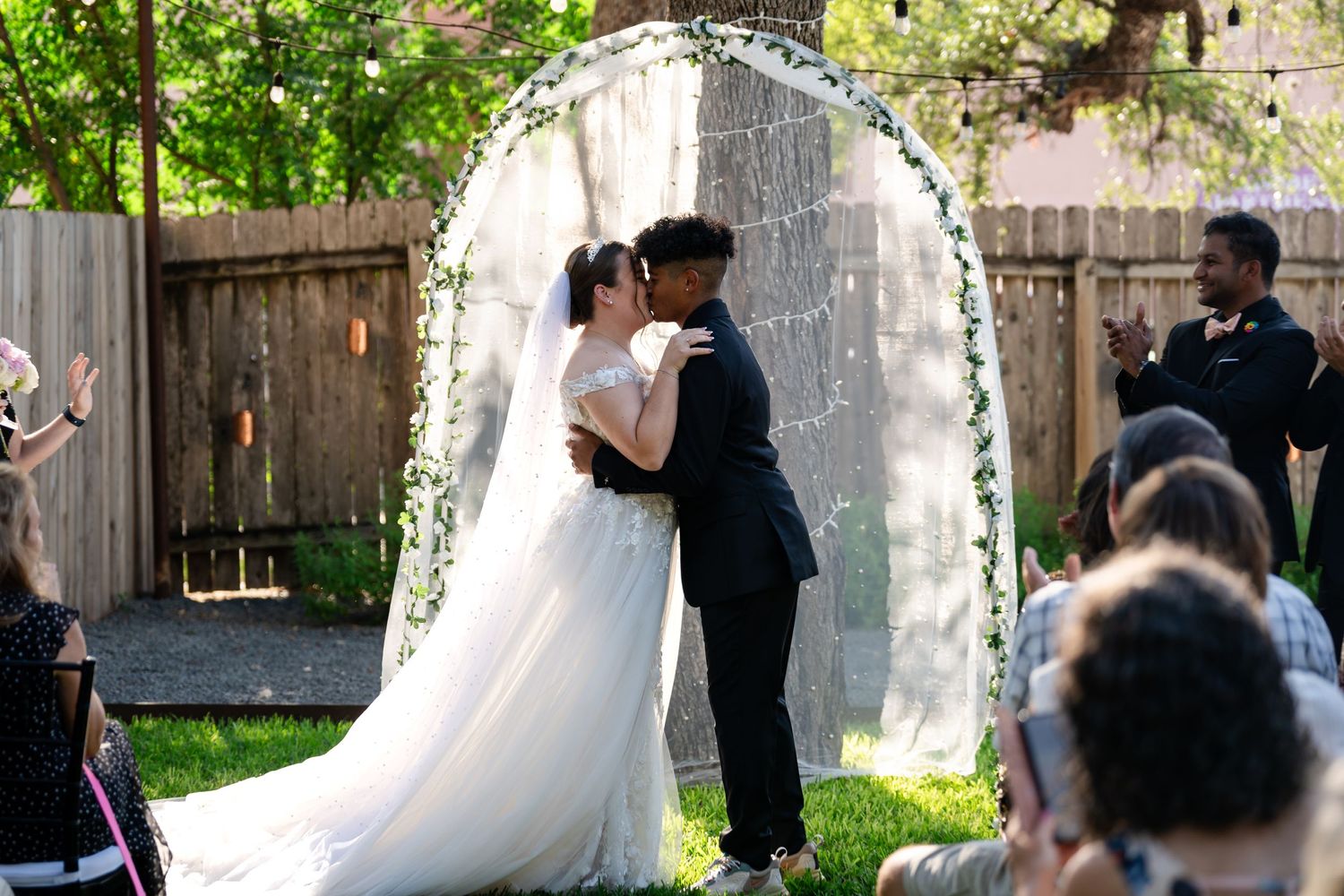 Newlyweds share their first kiss under a white floral arch as guests look on.