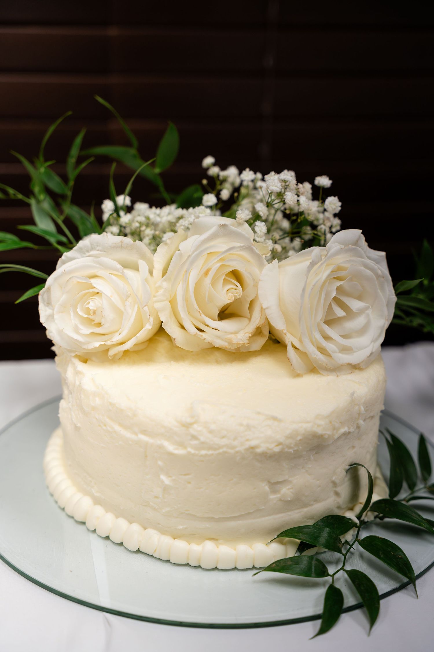 Elegant white buttercream cake decorated with fresh white roses, baby's breath and green foliage on a glass cake stand.