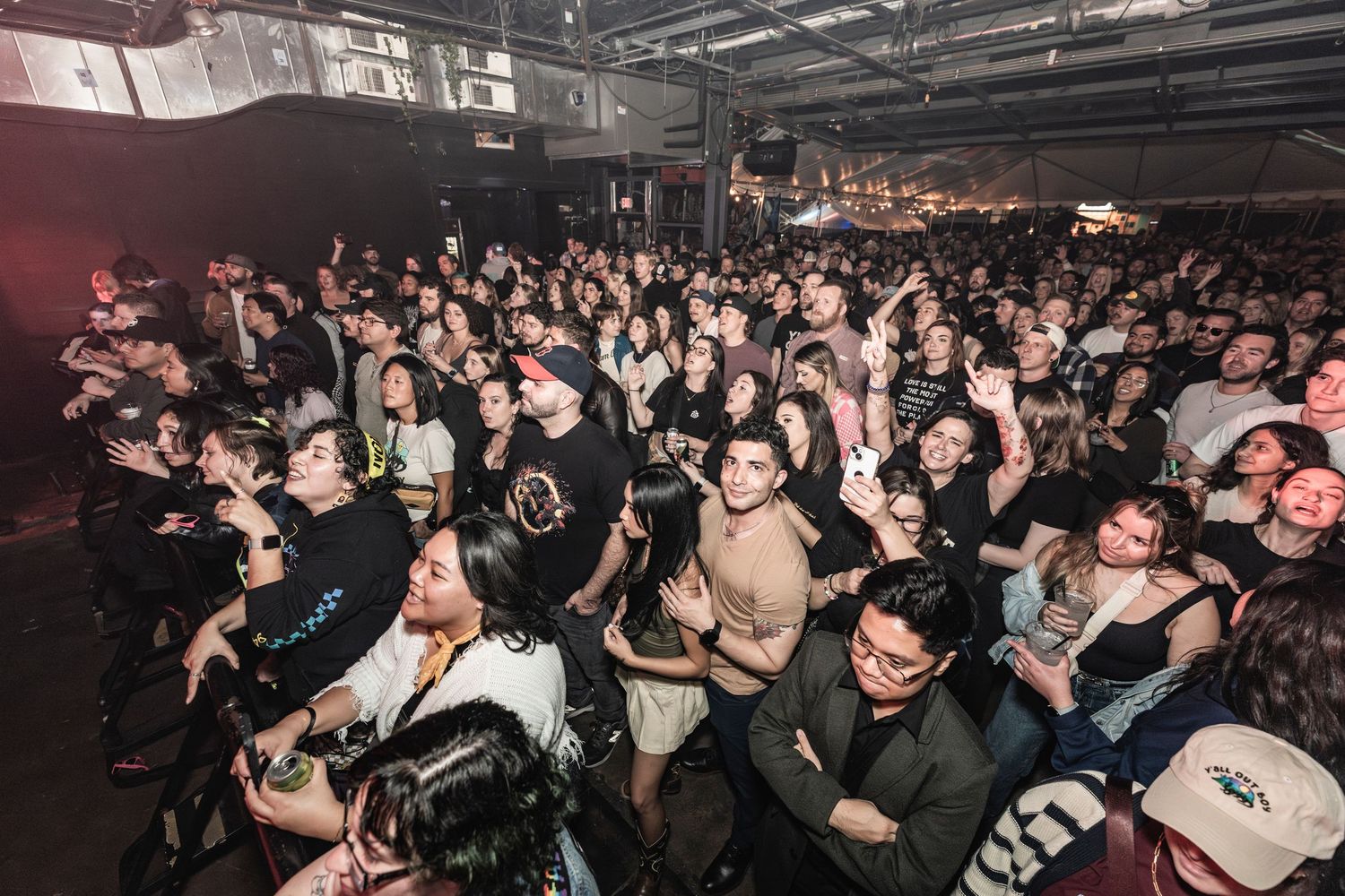 Large crowd of fans packed into a dark music venue during a live concert, with raised hands and energetic movement.