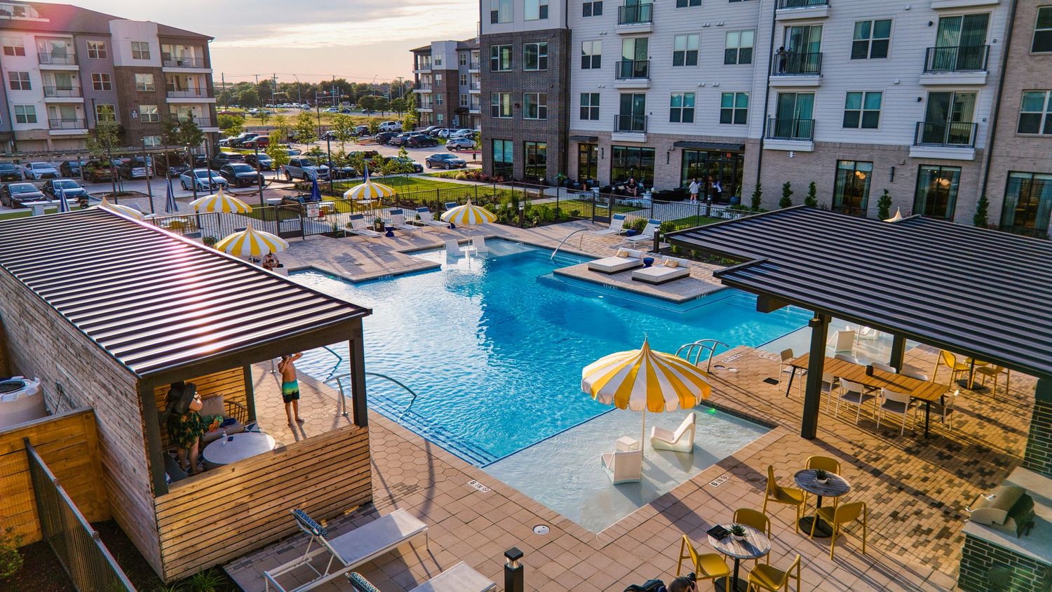 Sunset view of apartment complex pool area with wooden cabanas and yellow umbrellas.