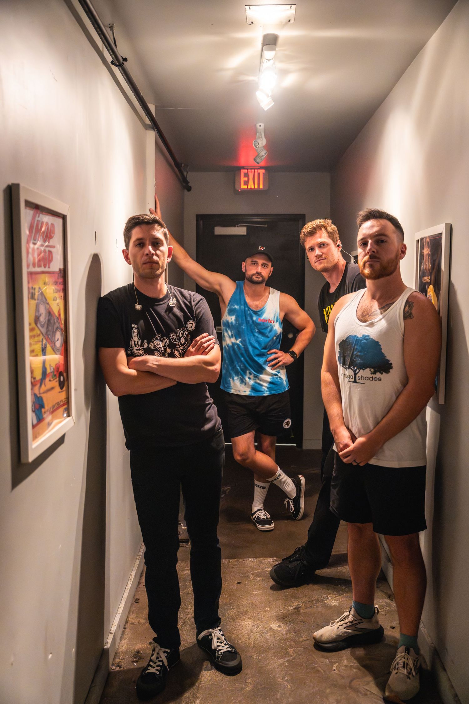 Four band members pose in a narrow hallway with exit sign and concert posters.